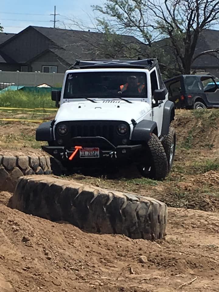 A white Jeep Wrangler navigates an off-road course over large tires in a dirt field.