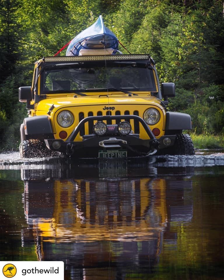 A bright yellow Jeep drives through shallow water in a forest, carrying a blue kayak on its roof rack.