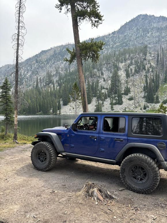 Blue off-road Jeep parked by a mountain lake with pine trees and rocky hills in the background
