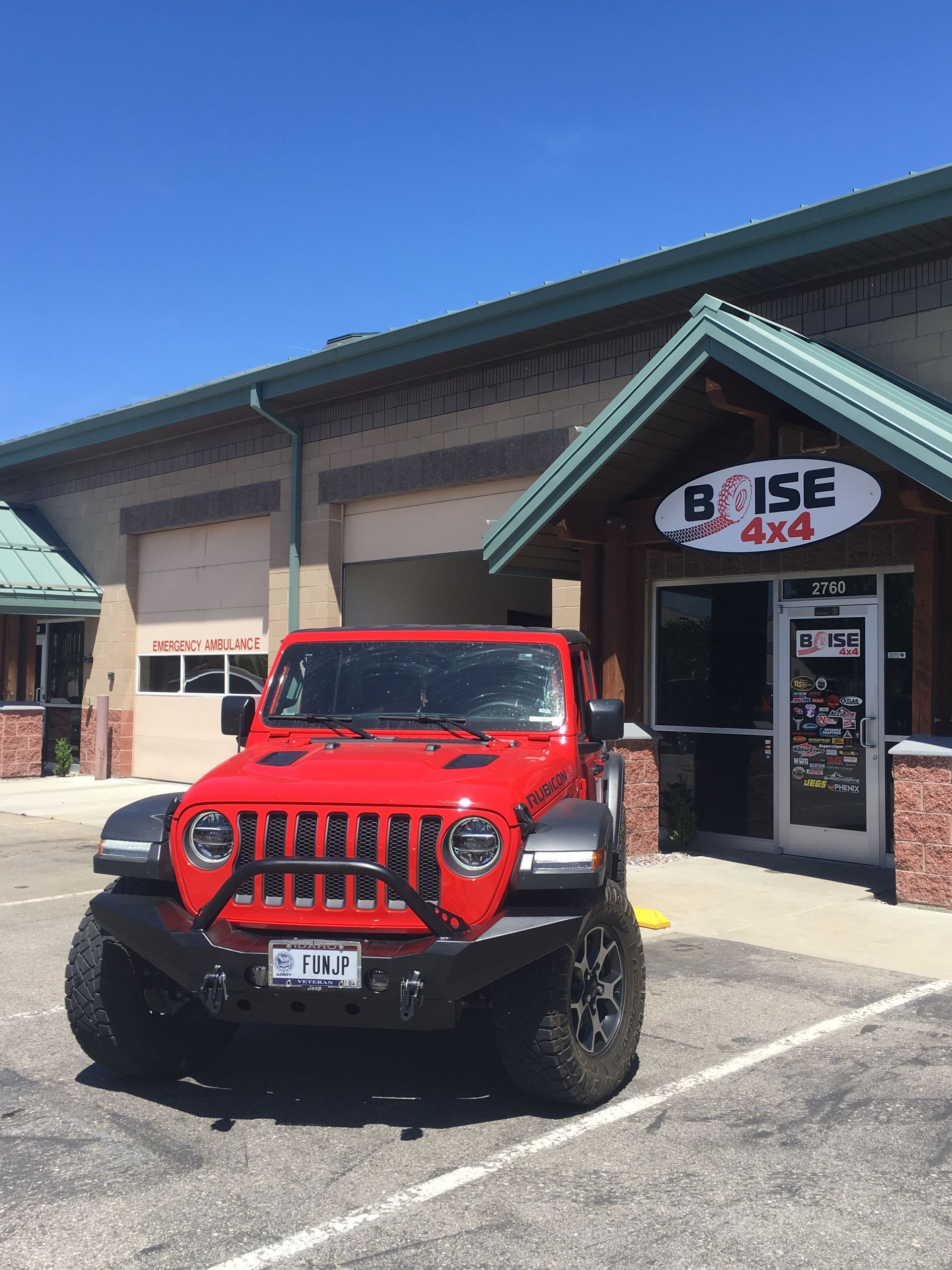 A bright red Jeep with a black aftermarket bumper parked in front of the Boise 4x4 shop under a clear blue sky.