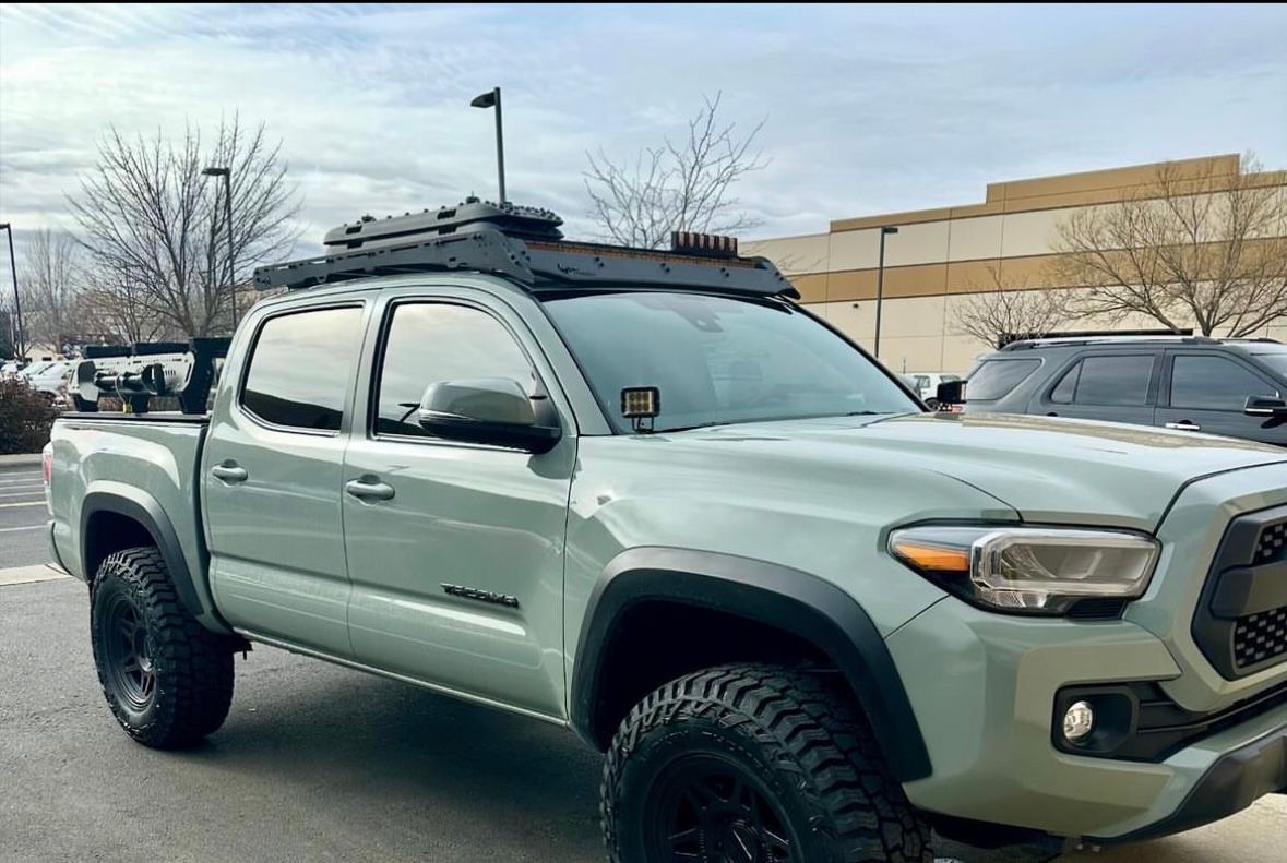 A lunar rock Toyota Tacoma truck with an off-road roof rack and storage box, parked in a parking lot.