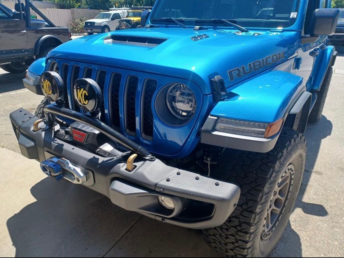 A bright blue Jeep Wrangler Rubicon parked outdoors, featuring a heavy-duty front bumper, winch, and KC auxiliary lights.