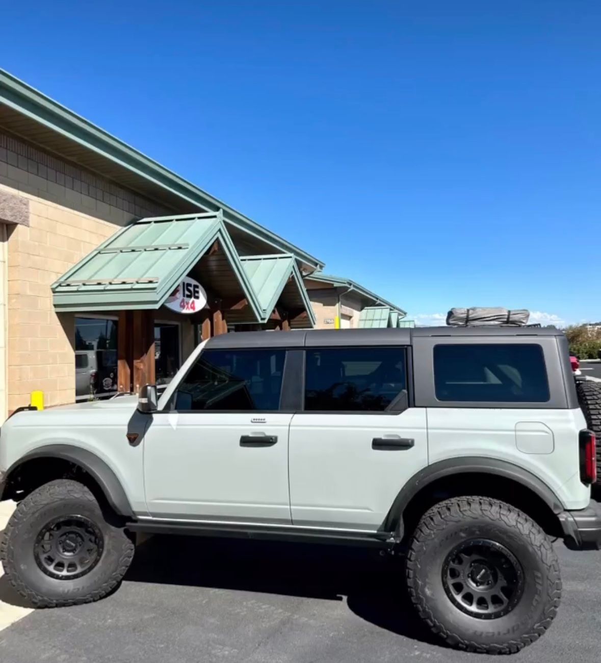 A light gray, four-door Ford Bronco with black wheels and a roof rack parked beside a building with a green awning.