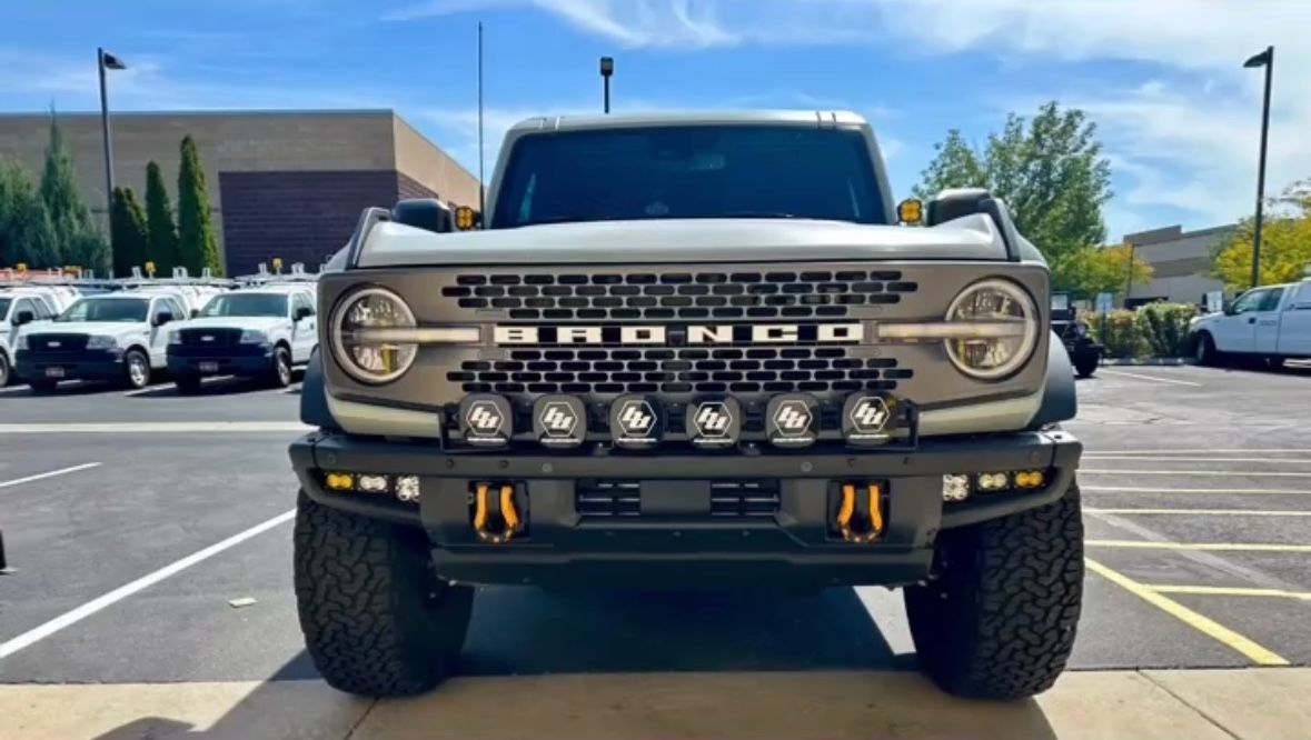 Front view of a gray Ford Bronco with off-road lights and orange tow hooks, parked in an outdoor lot.