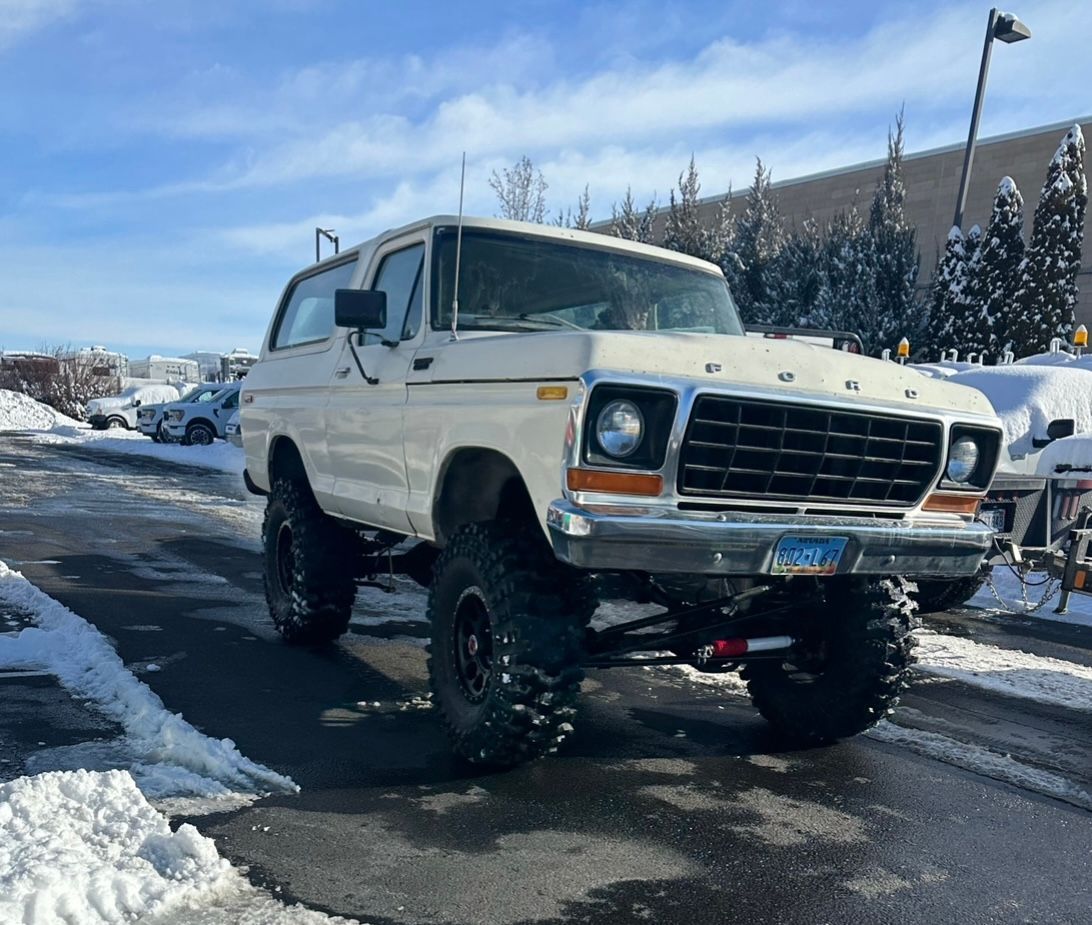 A white vintage Ford Bronco with large off-road tires parked on an asphalt lot covered in light snow.