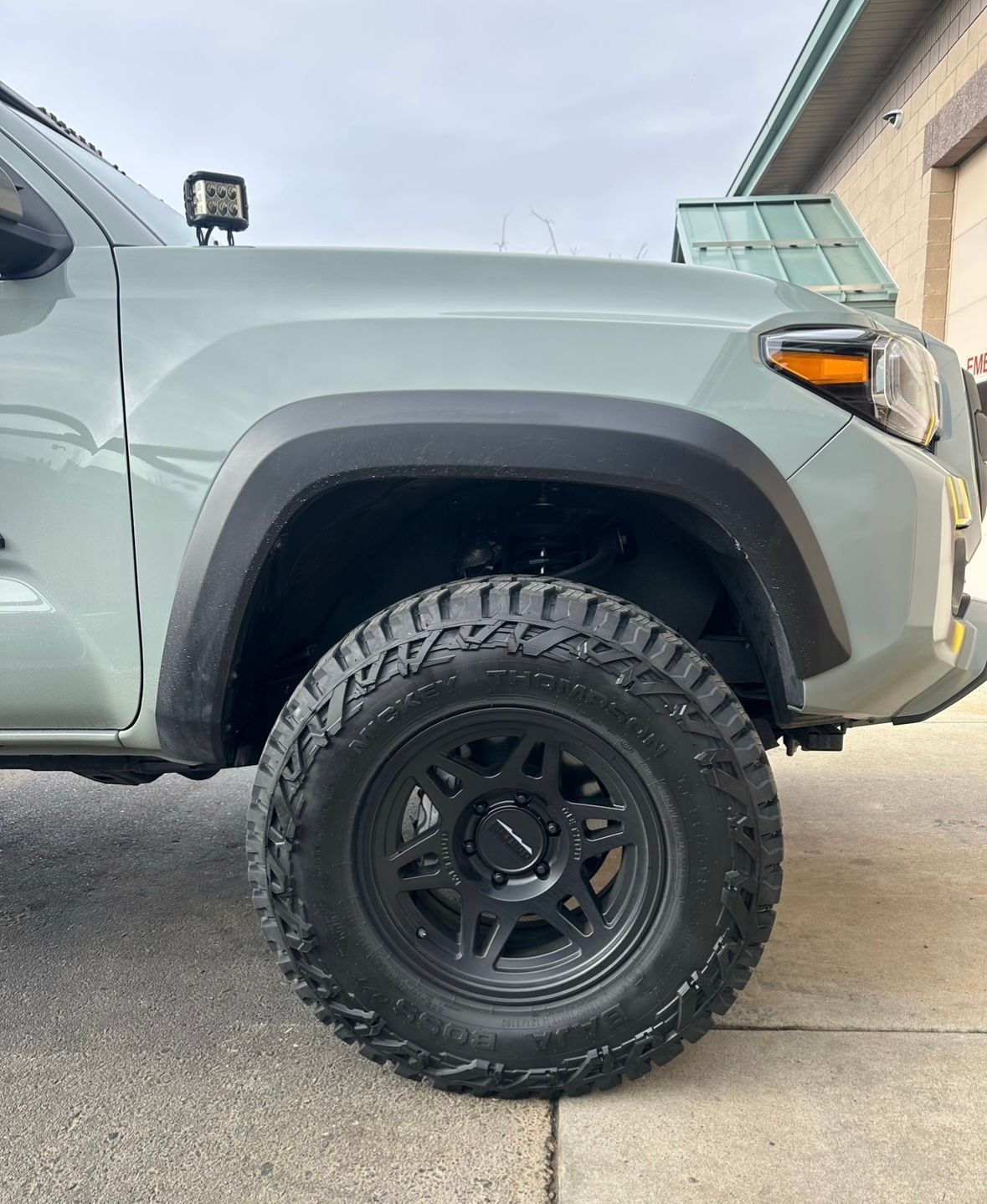 Close-up side view of a sage green Toyota Tacoma’s front fender, black off-road wheel, and knobby tire parked on concrete.