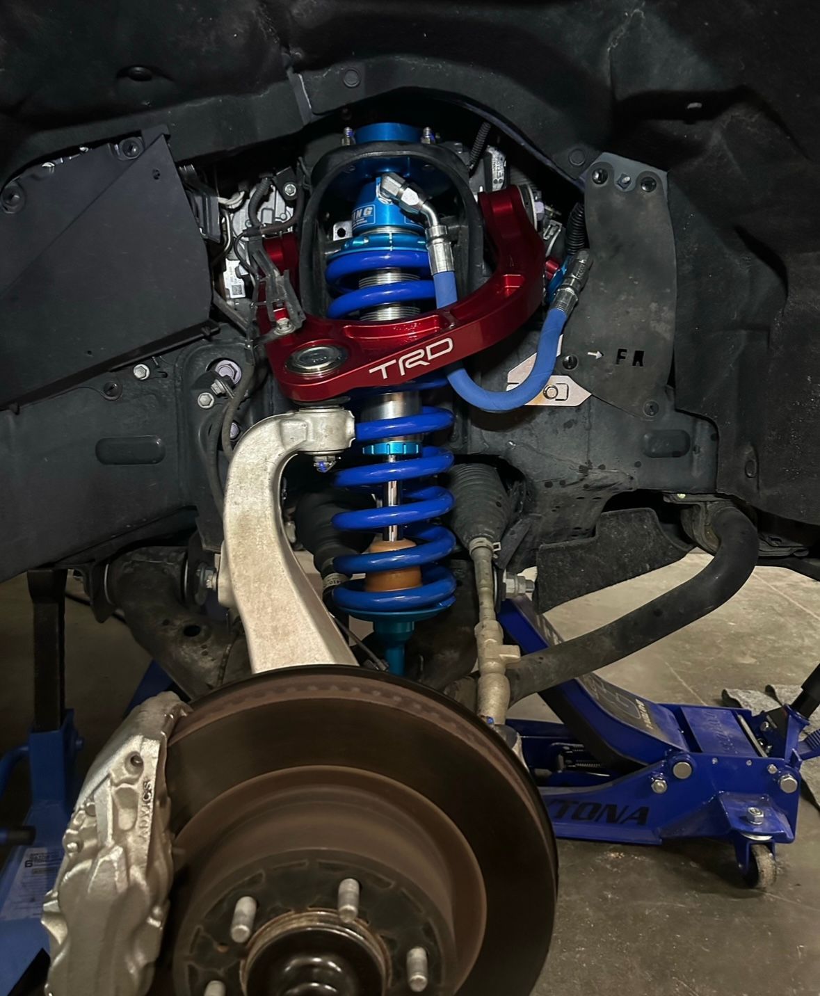 A close-up view of a vehicle's front suspension system, featuring blue coil springs and a red TRD control arm.