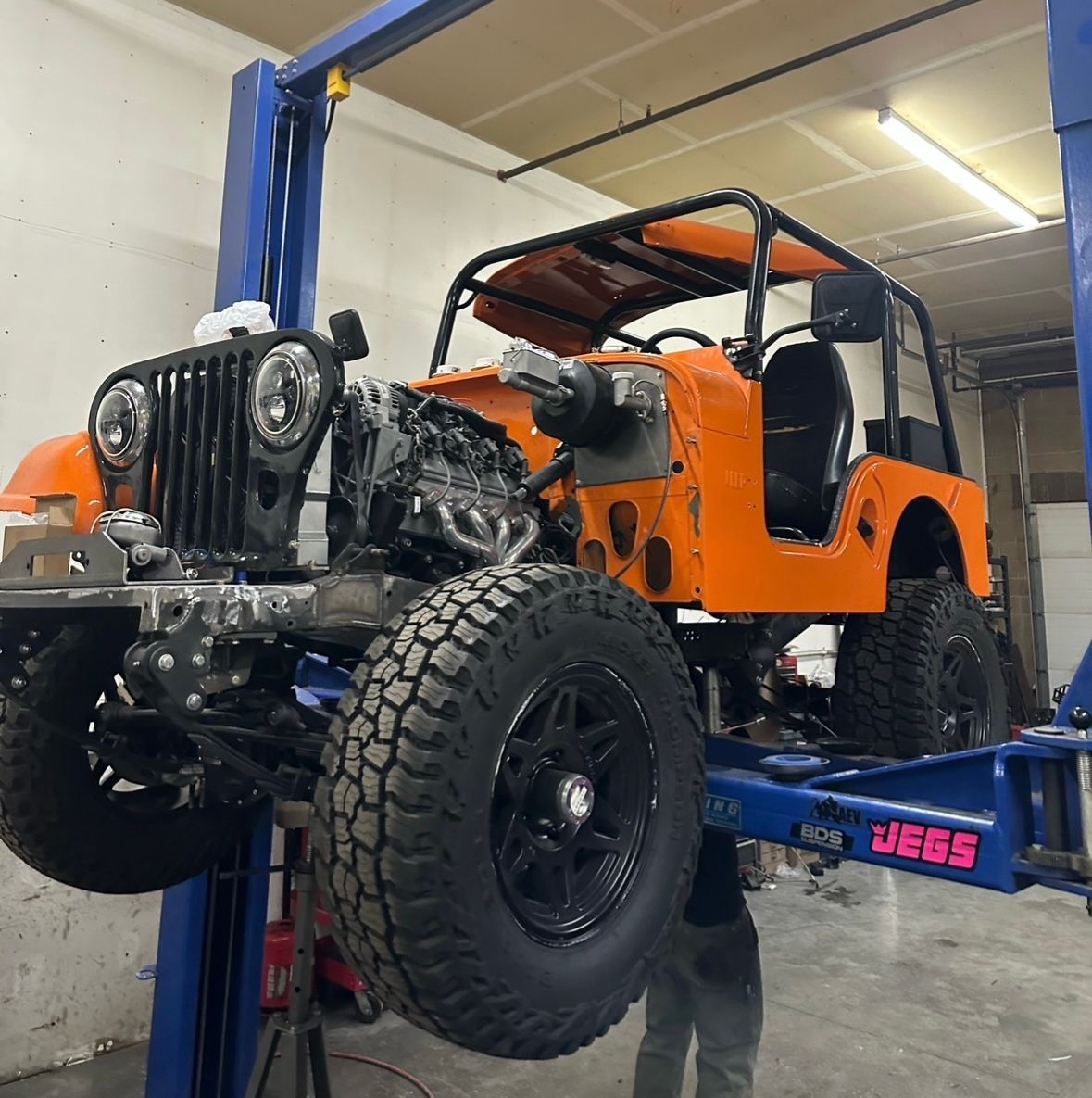 An orange Jeep with a black roll cage is raised on a blue vehicle lift in a garage.