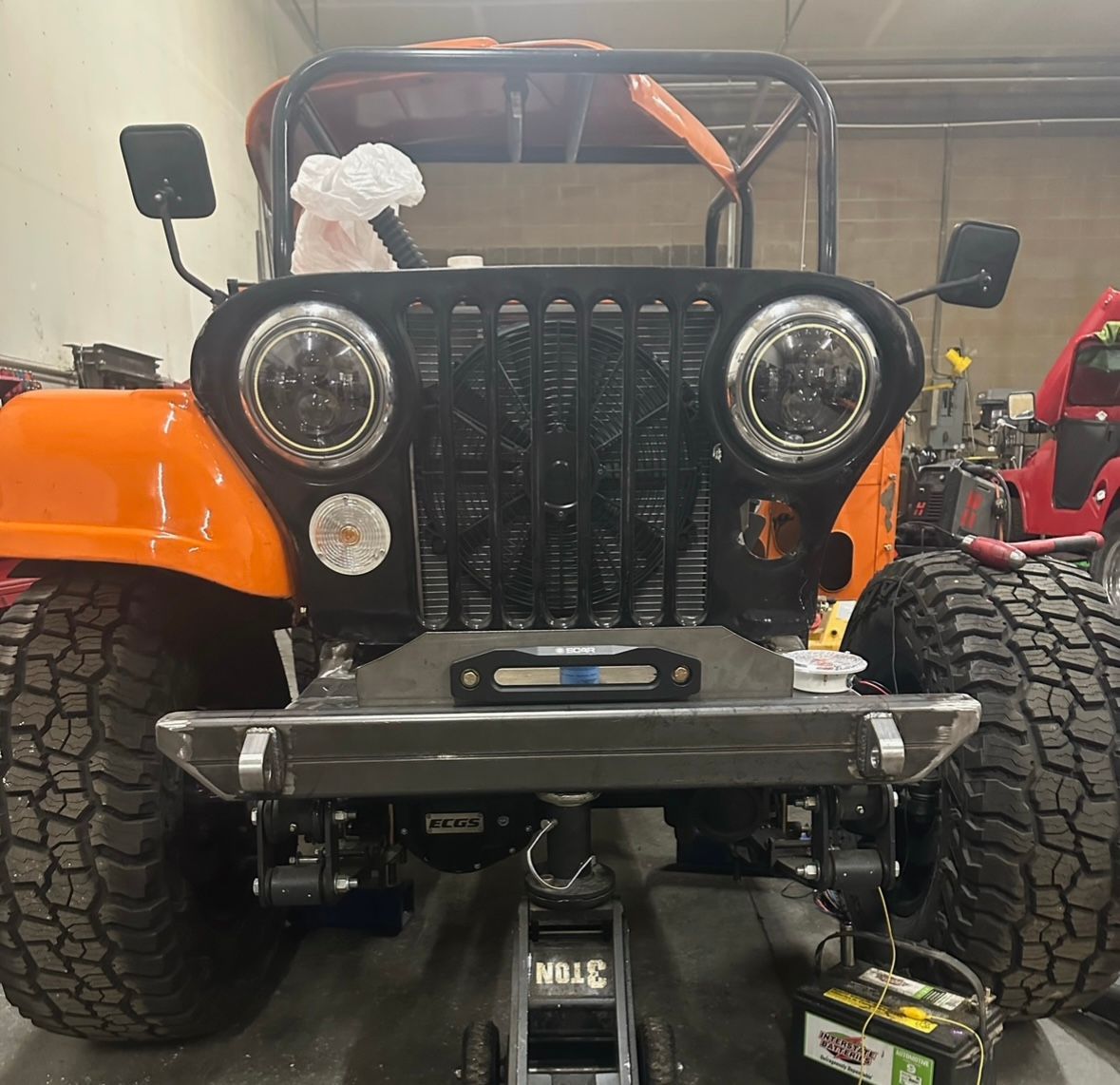 Front view of an orange Jeep undergoing maintenance in a garage, featuring a black grille, large tires, and a floor jack.