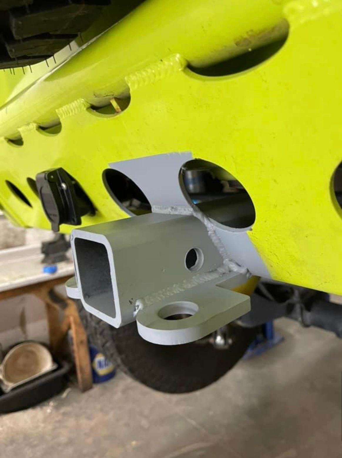 A grey trailer hitch receiver with a tow loop welded to a bright yellow vehicle bumper in a workshop setting.