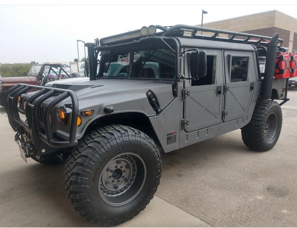 A gray Hummer H1 with off-road tires, a roof rack, and a front brush guard parked on an asphalt lot.
