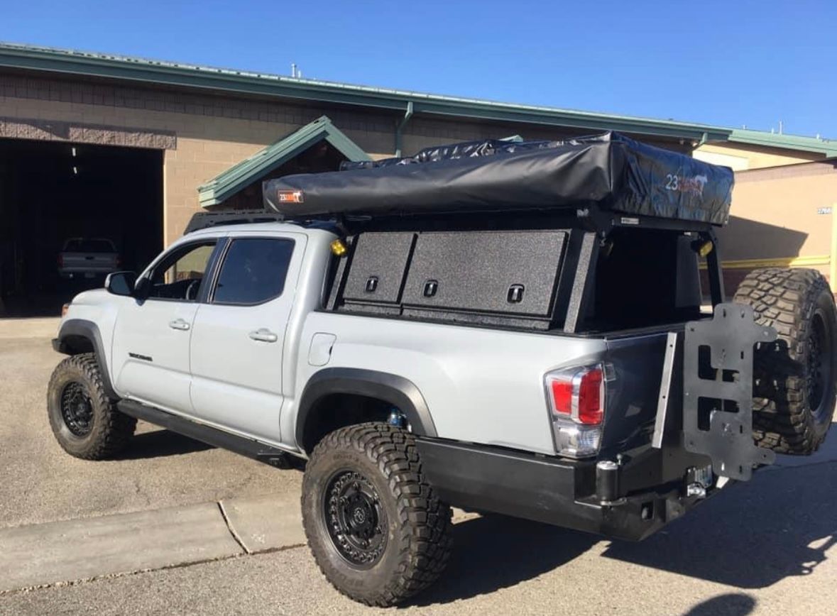 Silver Toyota Tacoma pickup truck with off-road tires, bed rack, and rooftop tent parked outside a building.