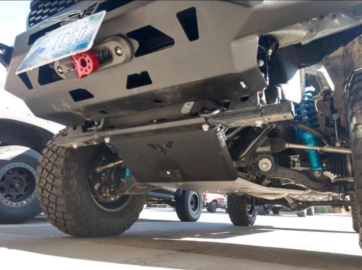 Low-angle view of a modified truck's undercarriage, featuring a black aftermarket bumper, skid plate, and blue suspension.