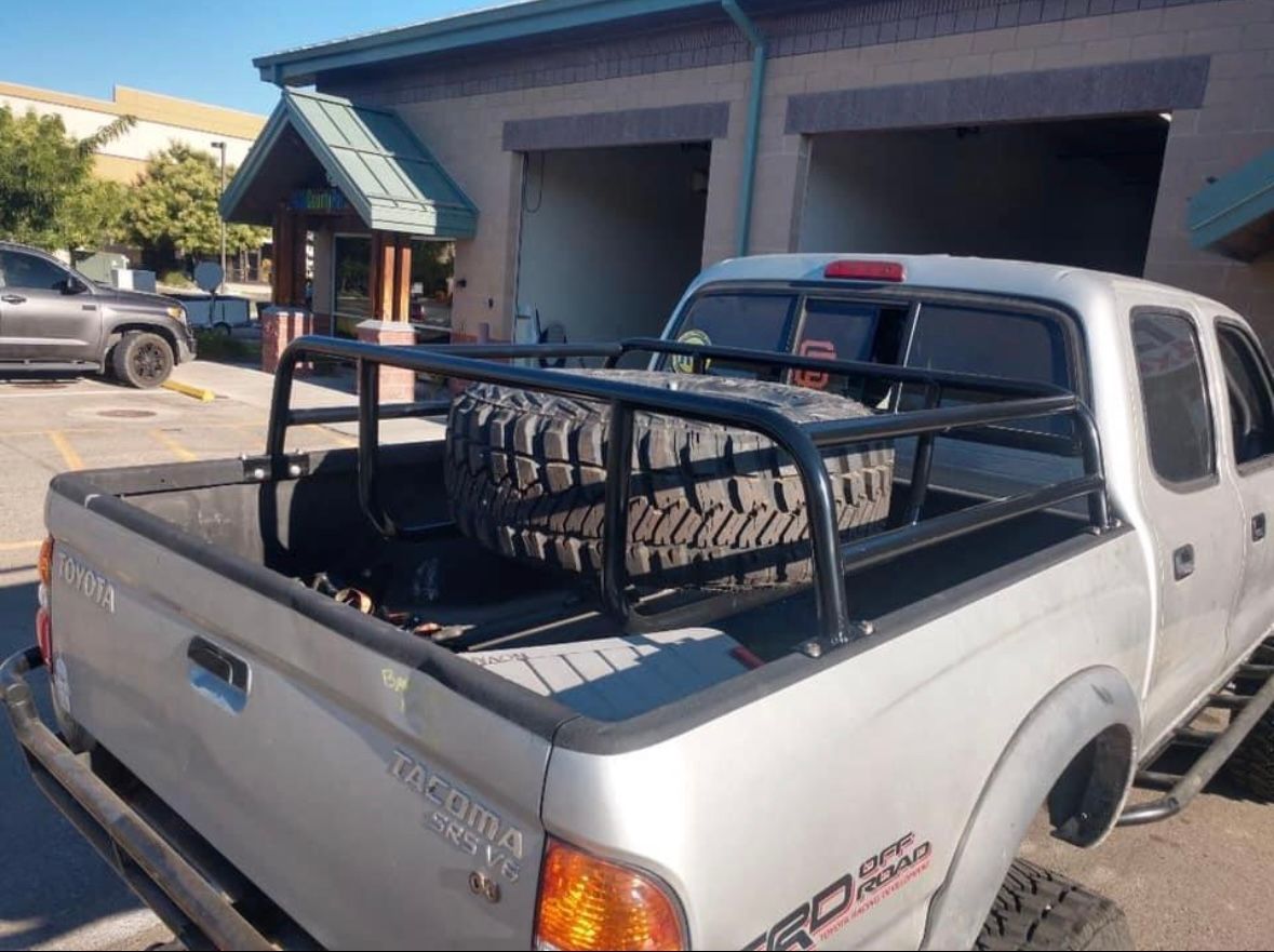 A silver Toyota Tacoma pickup truck with a large black bed rack carrying a spare off-road tire in a parking lot.