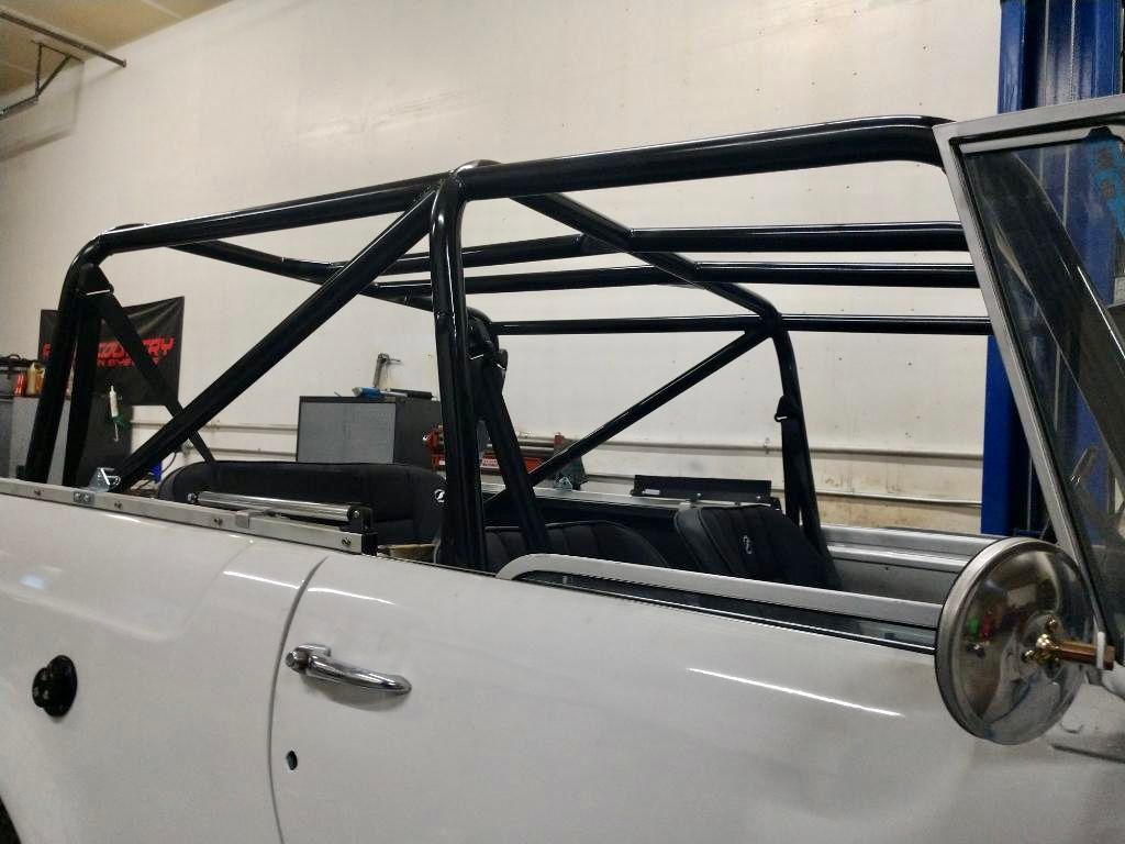 A side view of a white vintage vehicle featuring a custom-installed black tubular roll cage in an automotive workshop.