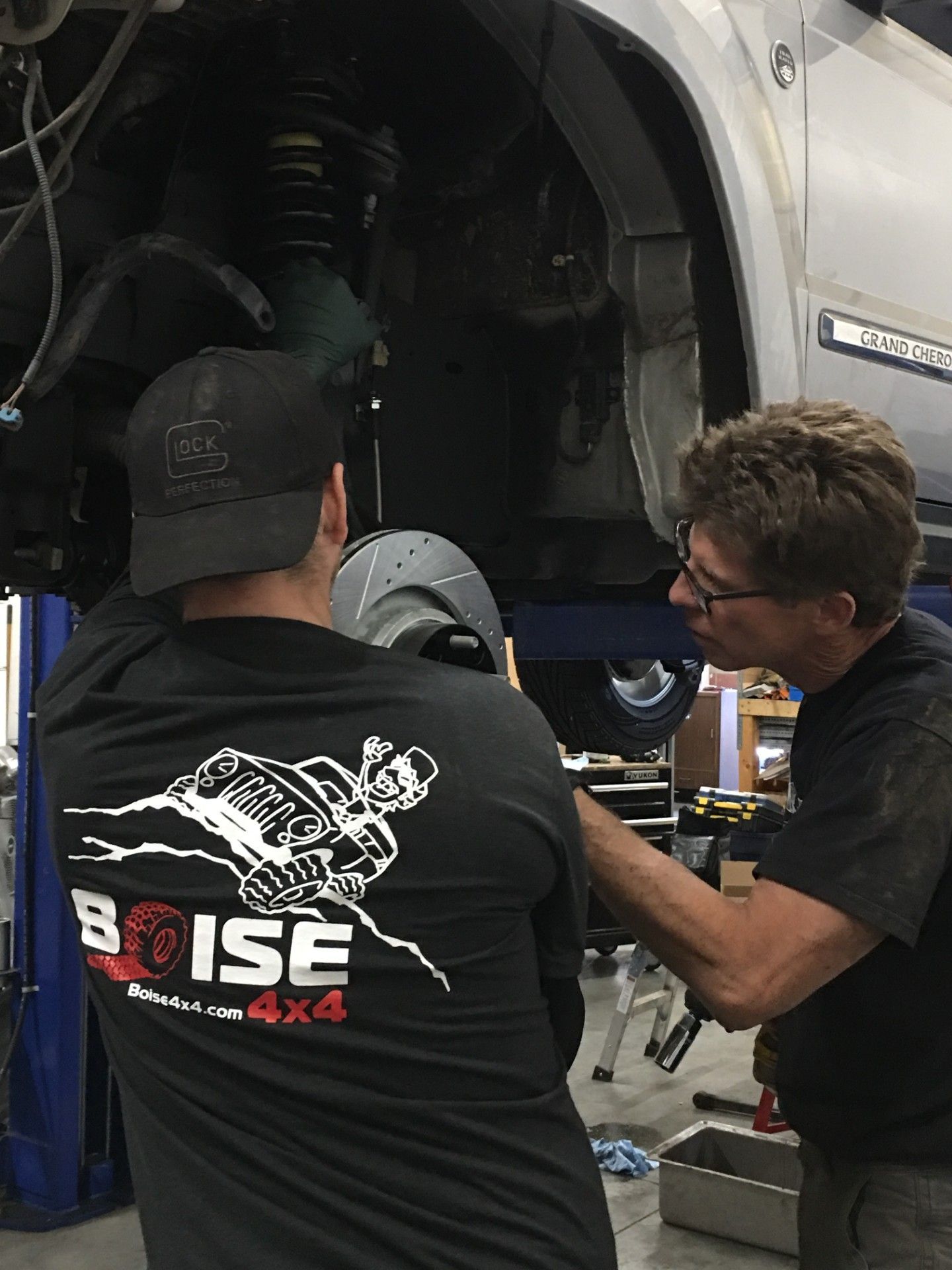 Two mechanics work on the suspension and brake system of a vehicle lifted in an auto repair shop.