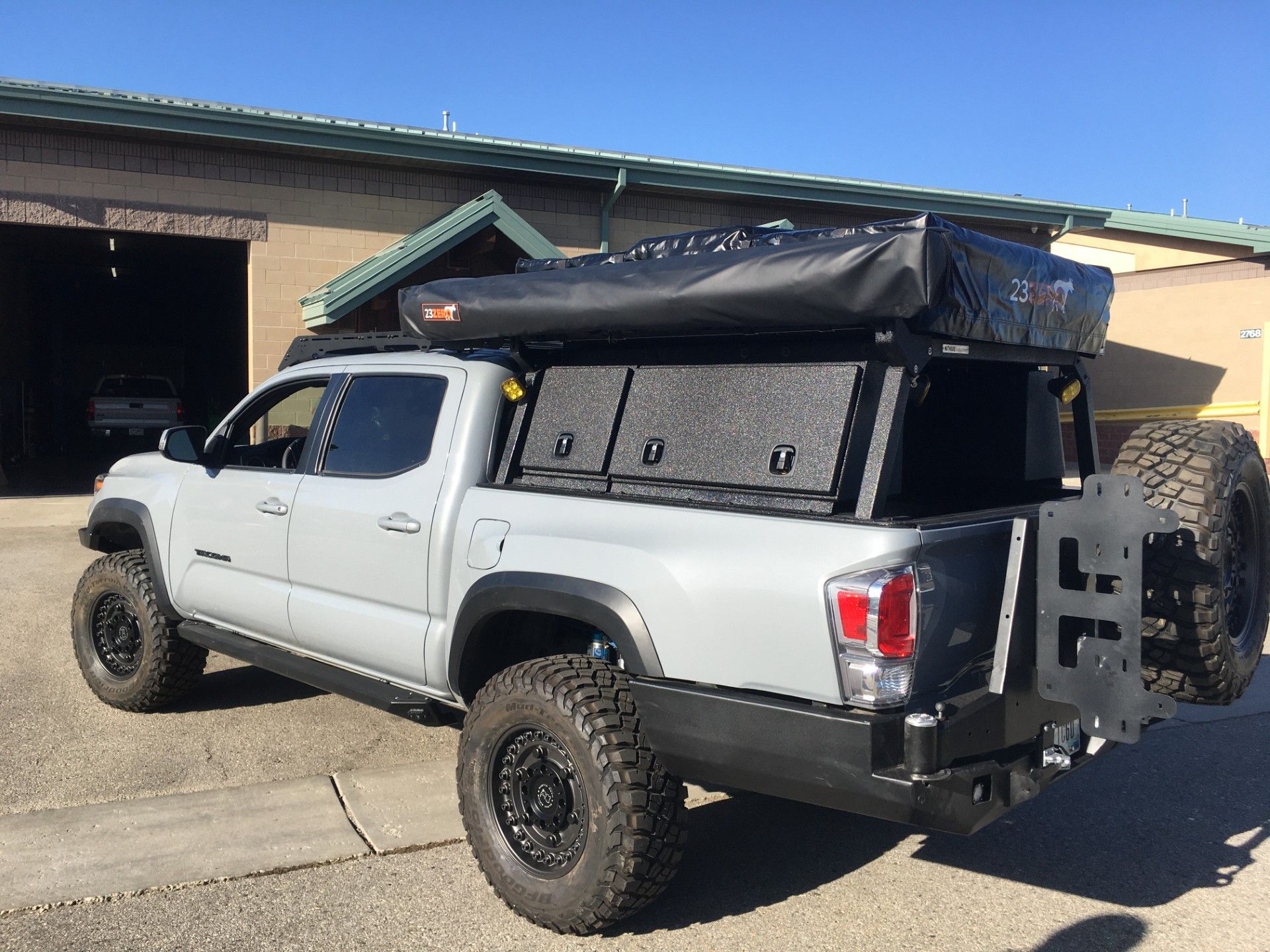 A gray pickup truck modified for off-roading, featuring a black roof tent, bed storage system, and rear spare tire carrier.