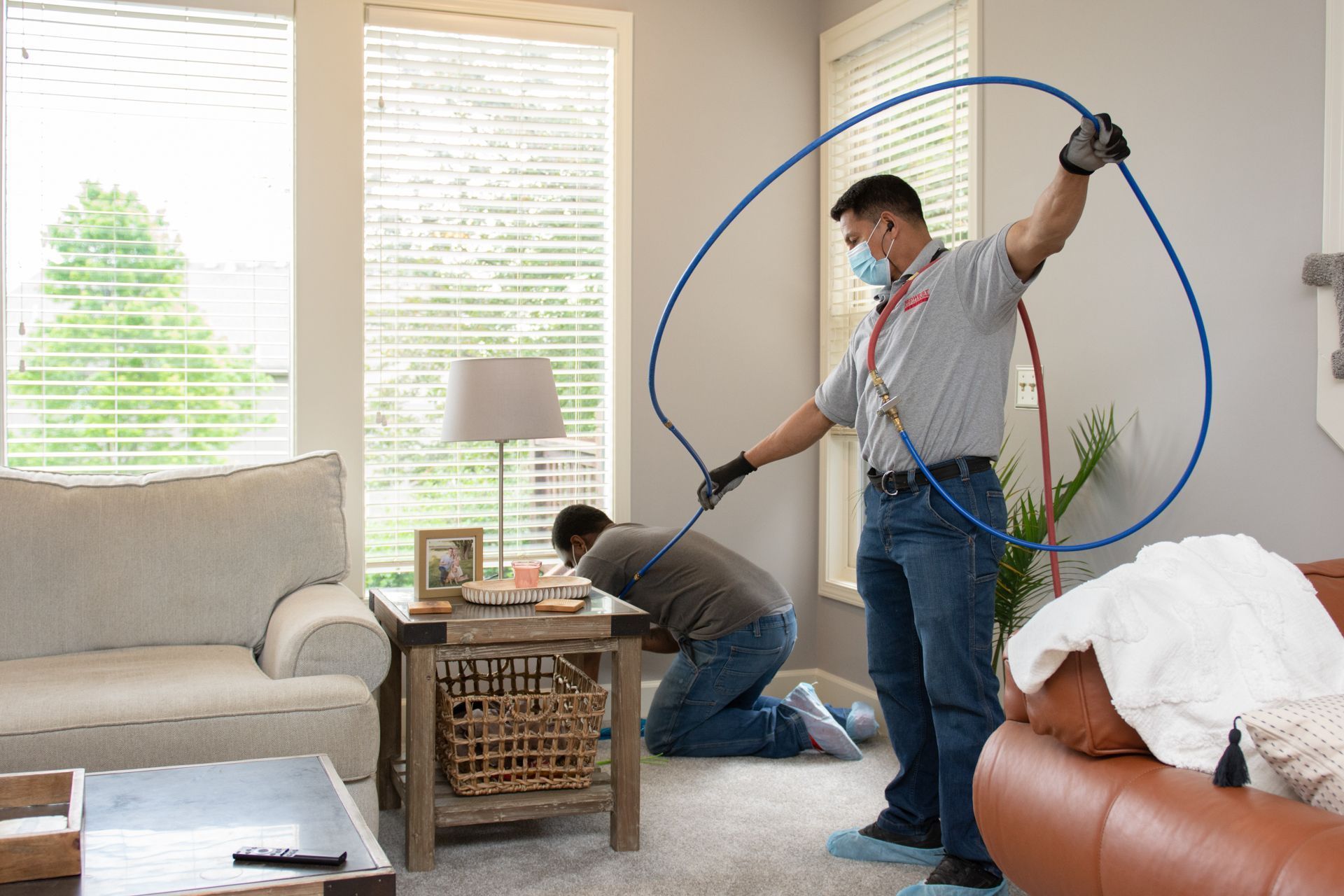 Two men in a living room, one using a hose on a wall, another looking at a table.