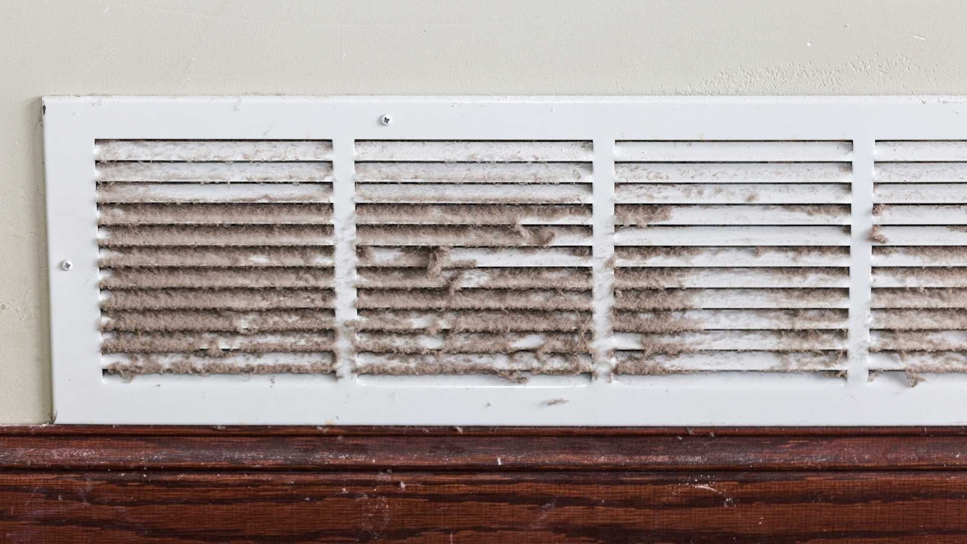 Dusty white air vent on a wall above a wooden baseboard.