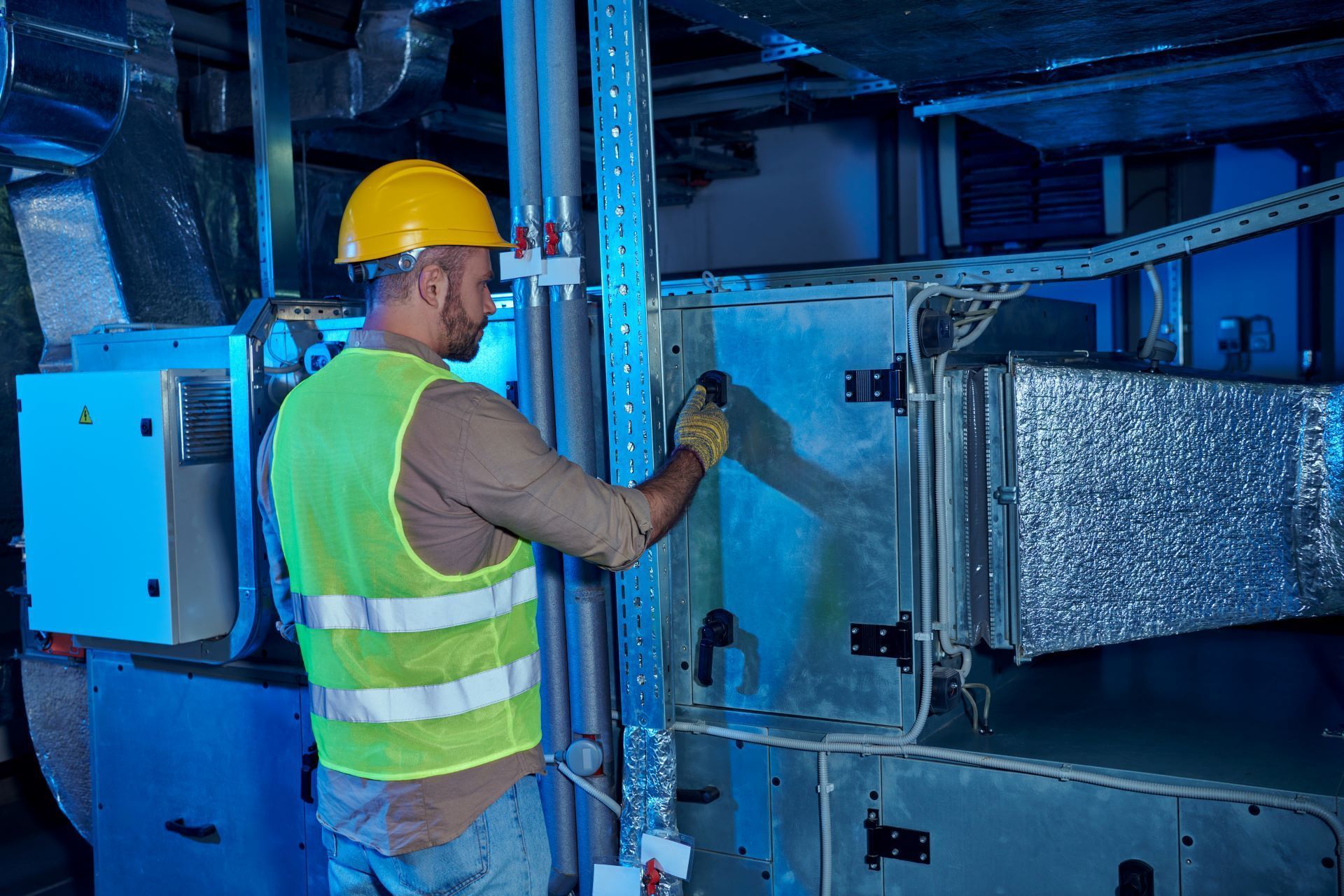 HVAC technician in hard hat and vest works on machinery in an industrial setting.