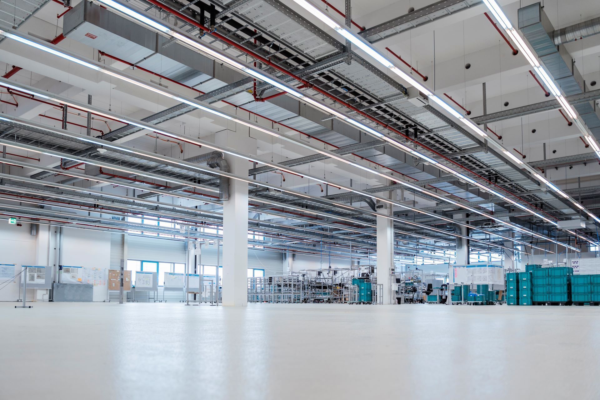 Large, bright factory interior; white floor, overhead pipes, machinery in the distance.