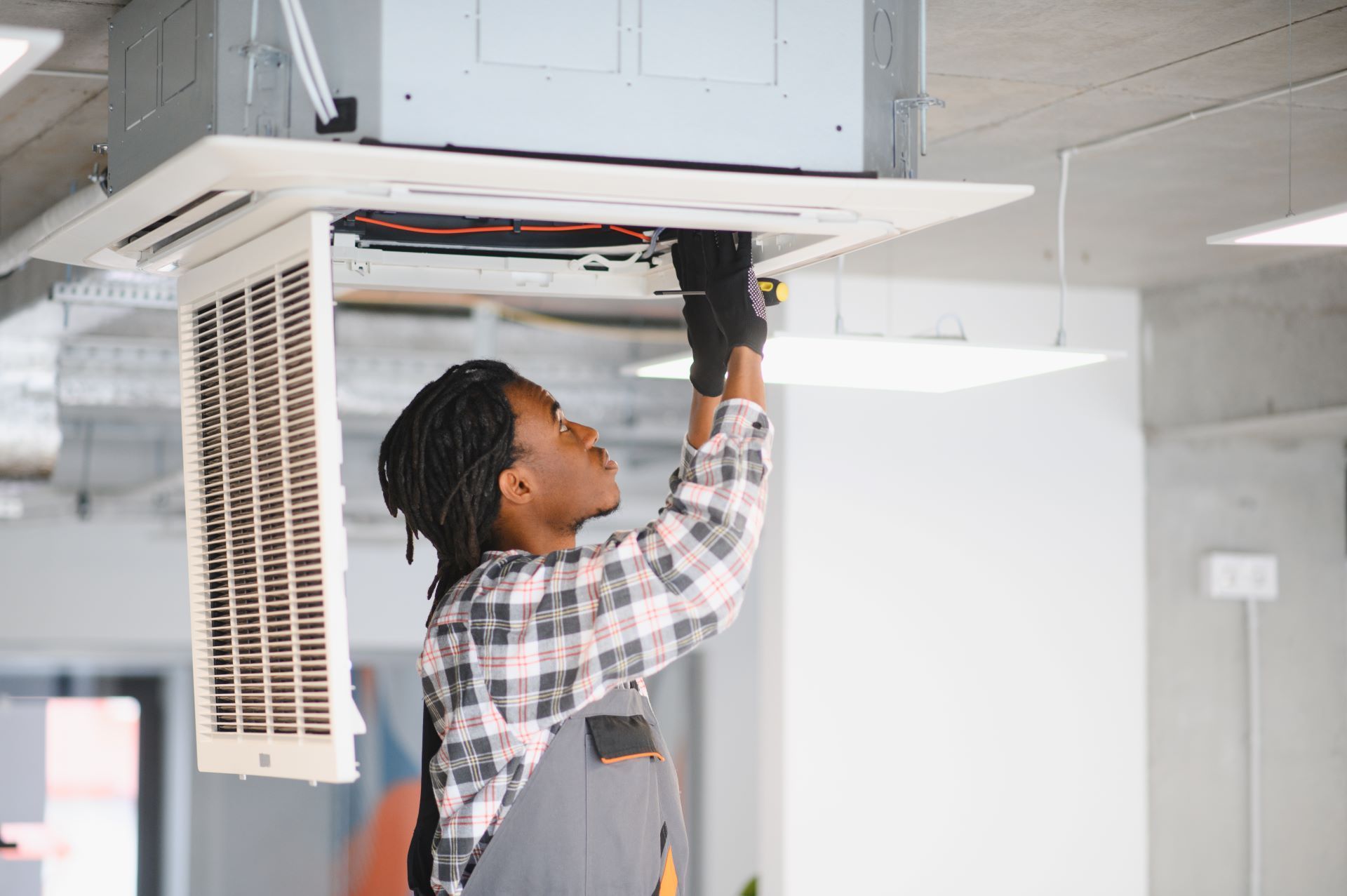 Black woman in overalls, fixing an air conditioning unit on a ceiling.