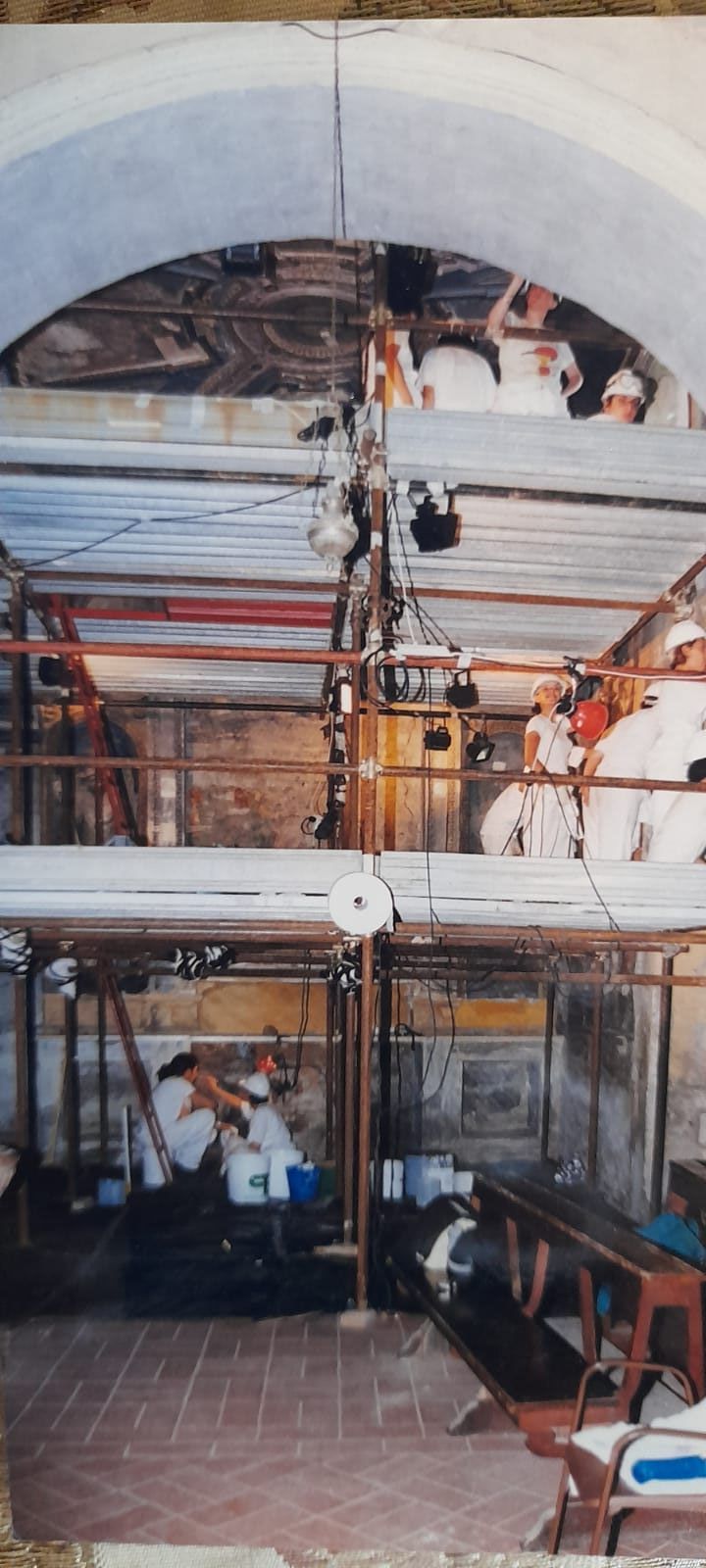 Vista interna di un'officina con impalcature, operai e macchinari. È visibile il soffitto a volta.