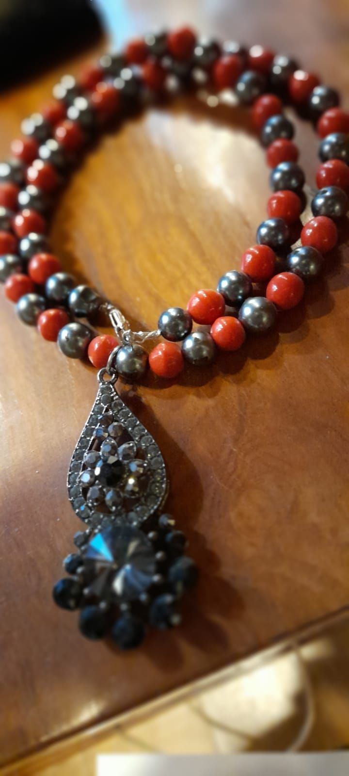 Collana con perline rosse e grigie, pendente a goccia con pietre nere, su una superficie di legno.
