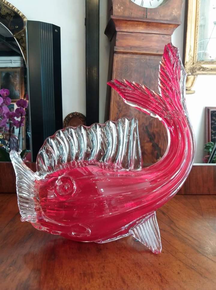 Scultura di pesce in vetro rosso su un tavolo di legno, con un orologio a pendolo e uno specchio sullo sfondo.