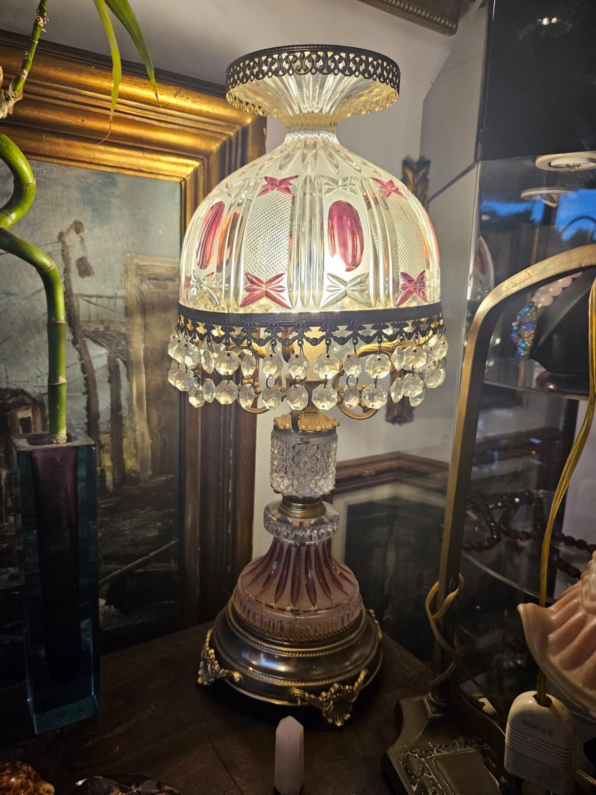 Lampada da tavolo in vetro decorato con frange di cristallo e dettagli rosa, appoggiata su legno scuro vicino a uno specchio e a un quadro.