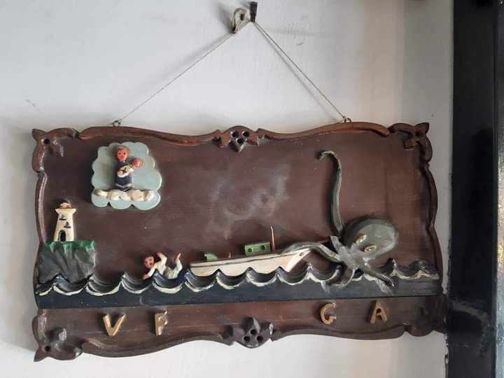 Scena nautica intagliata in legno con barca, polpo, faro e persone tra le onde; cartello appeso.