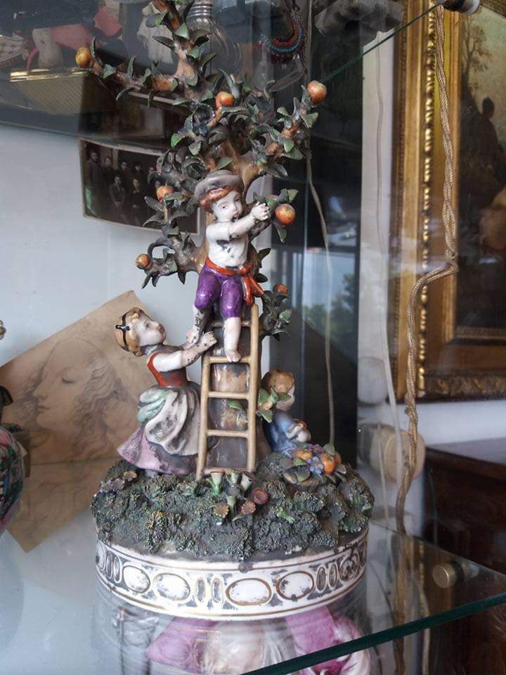 Statuina in porcellana raffigurante bambini che raccolgono frutta da un albero, su una base decorativa.