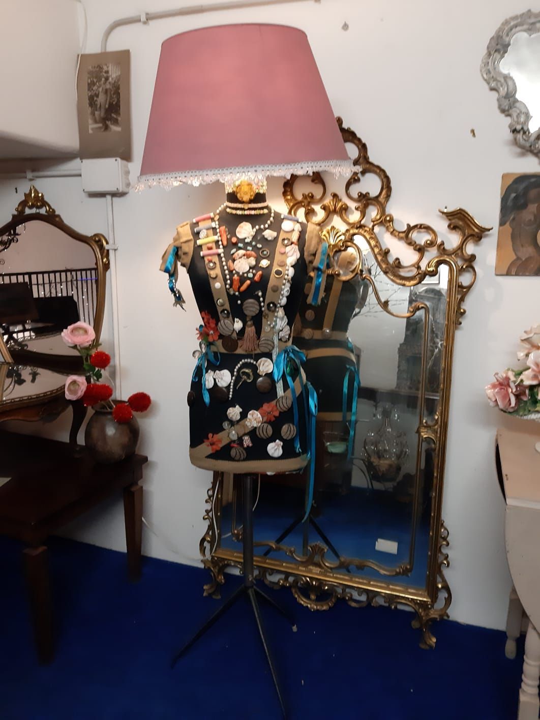 Una lampada a forma di manichino con paralume rosa è posta davanti a uno specchio con cornice dorata. Il manichino è decorato con varie decorazioni.