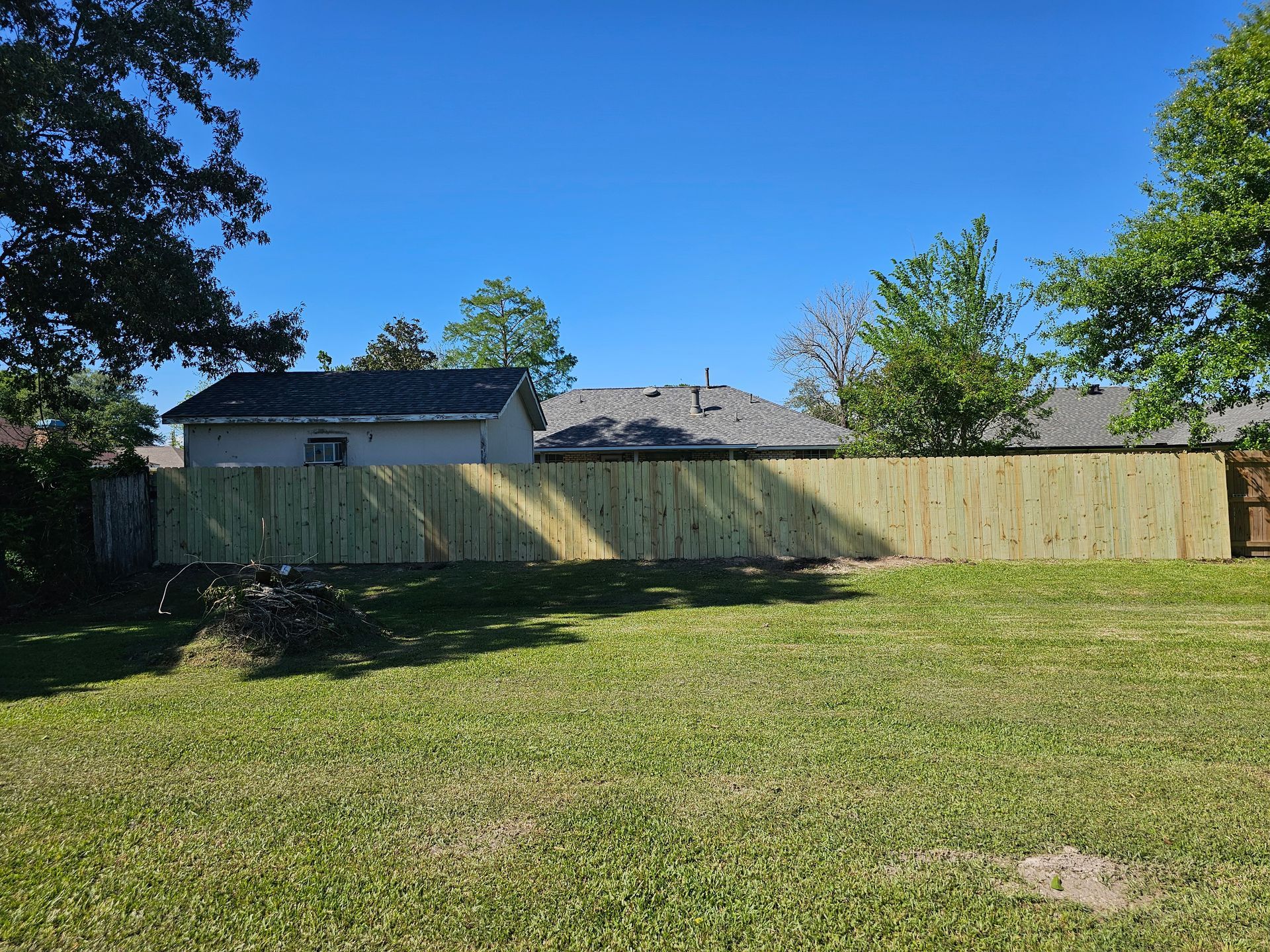 Backyard with a wooden fence, green lawn, and trees under a bright blue sky