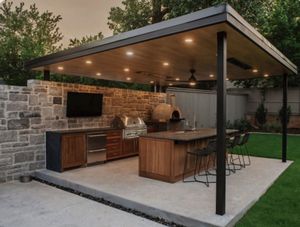 Outdoor stone patio with built-in stainless steel grill and cabinets beside a house, backed by trees and fencing