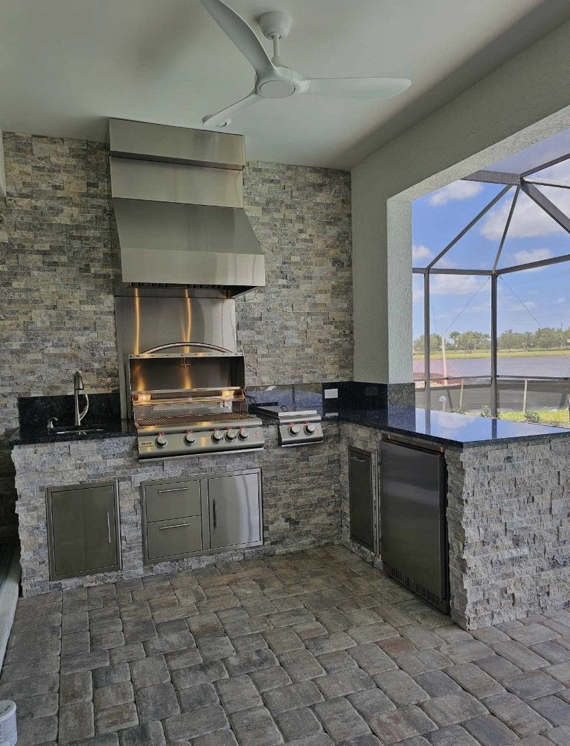 Outdoor stone kitchen with built-in grill, counter space, and screened view of a pool and lake.