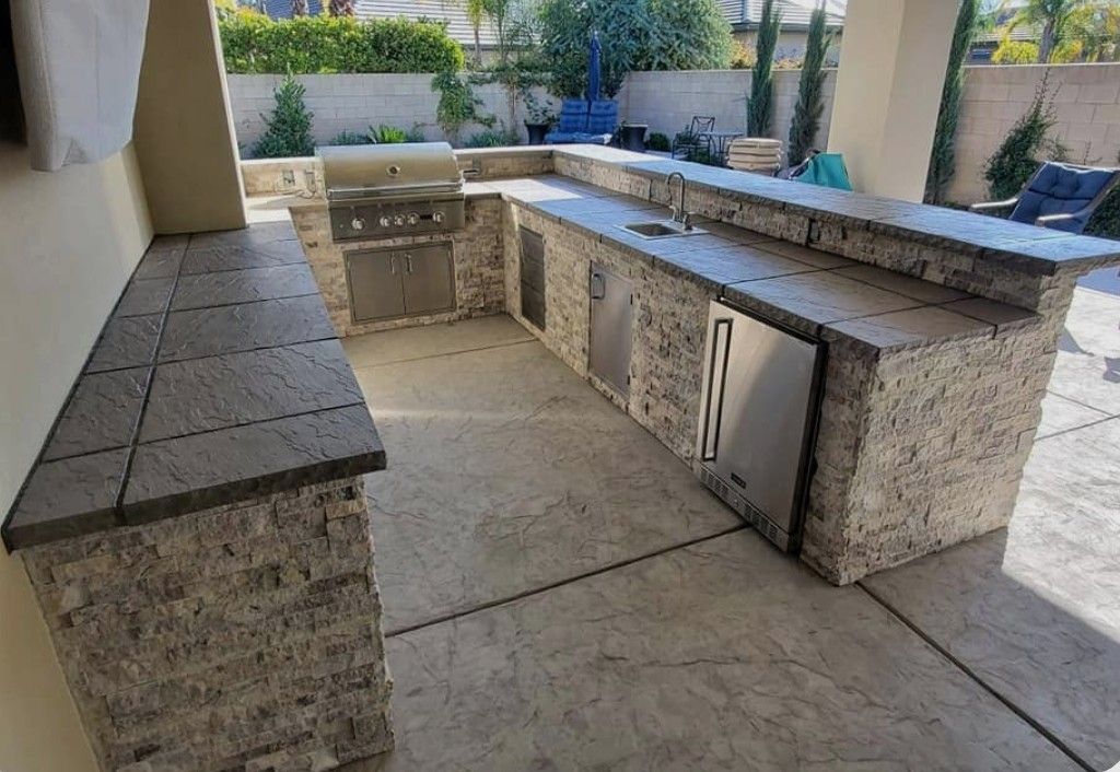 Outdoor stone barbecue and counter area under a covered patio, with built-in cabinets and seating benches.