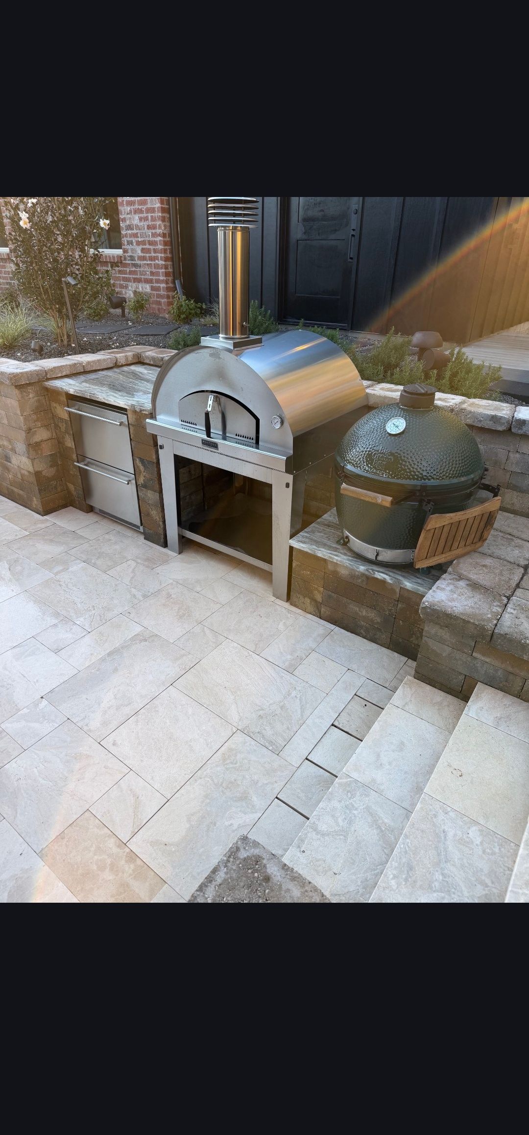 Stone outdoor pizza oven with stacked firewood beside a tiled patio and brick wall.