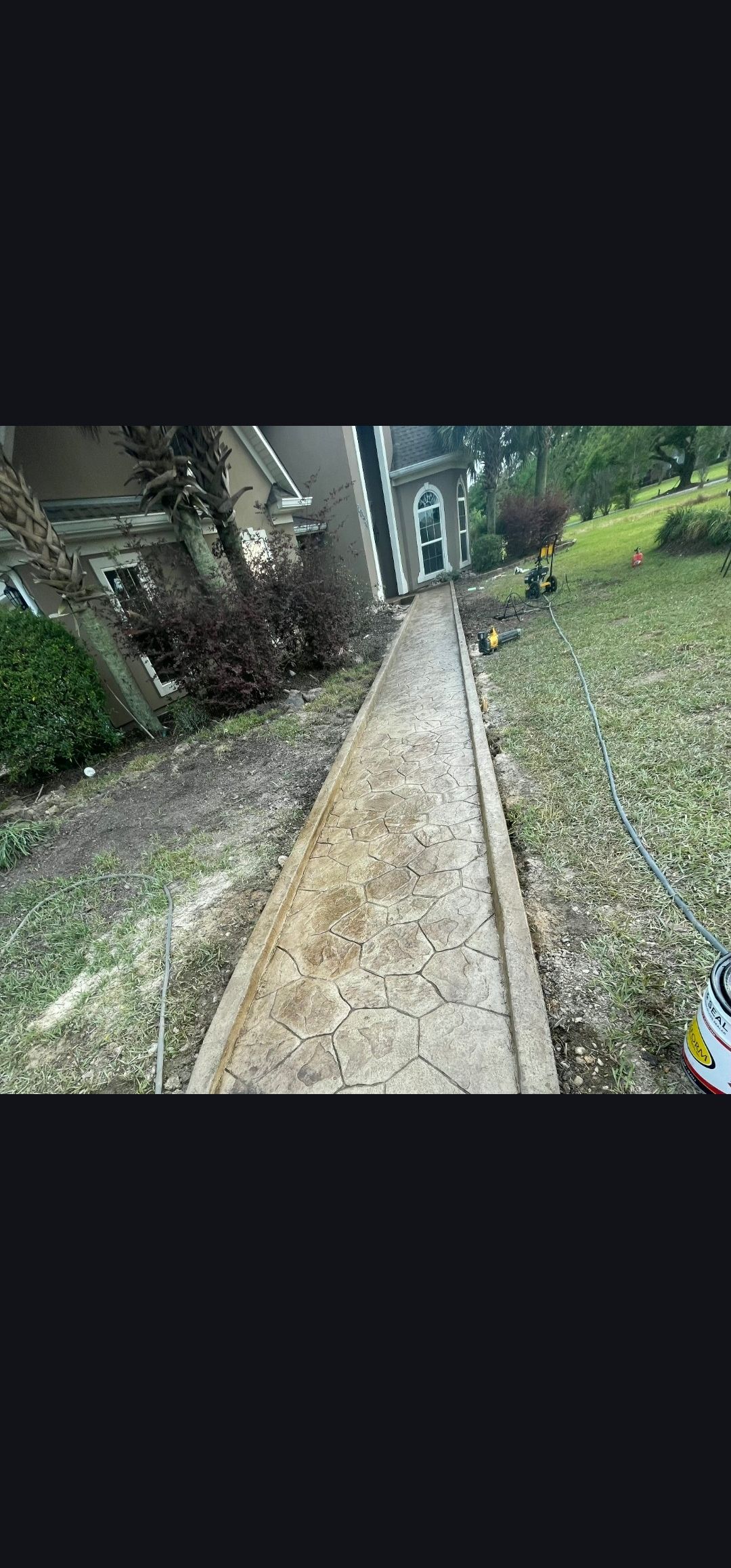 Narrow concrete path beside a grassy yard with trees and buildings in the distance