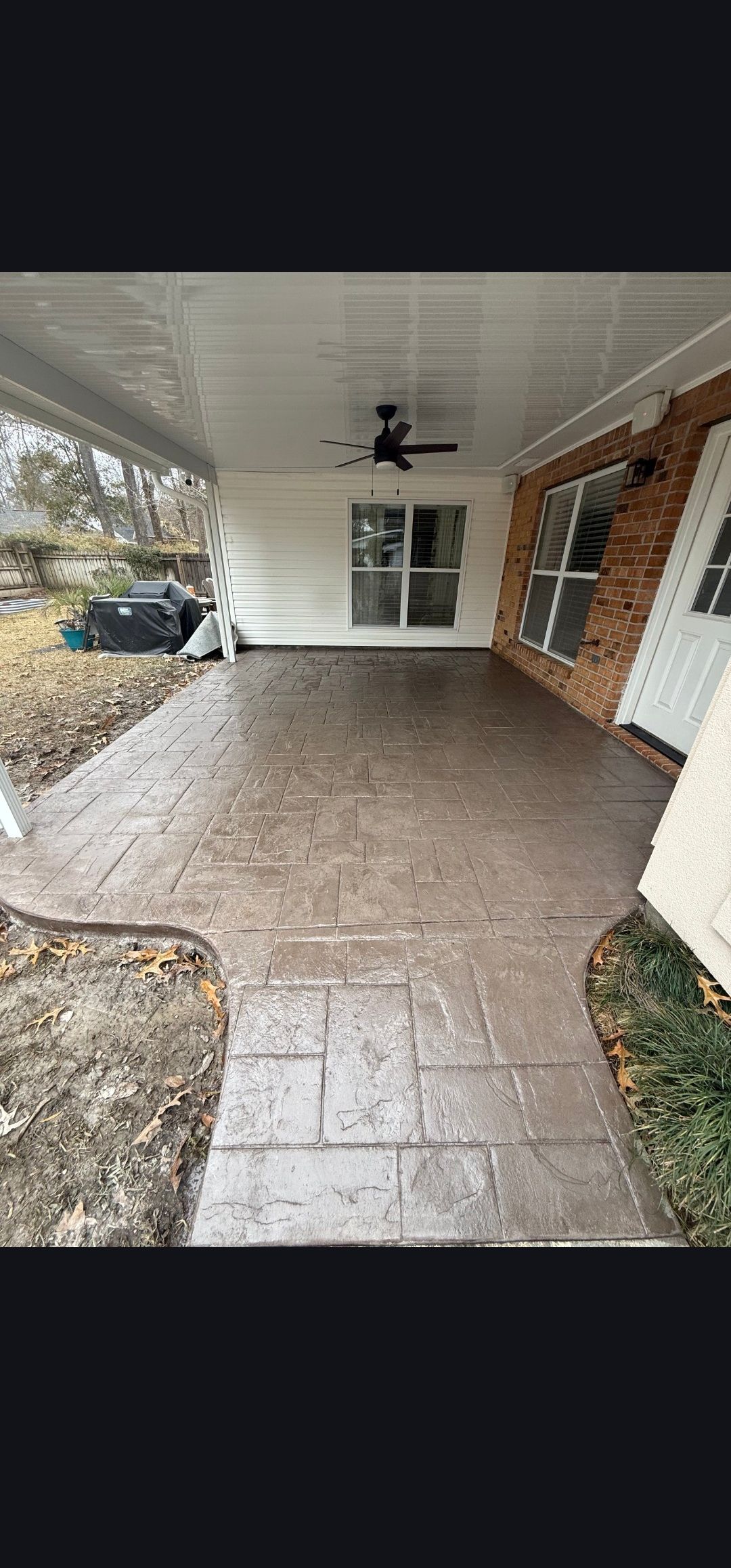 Covered brick patio with ceiling fan, white double doors, and brick wall beside a driveway.