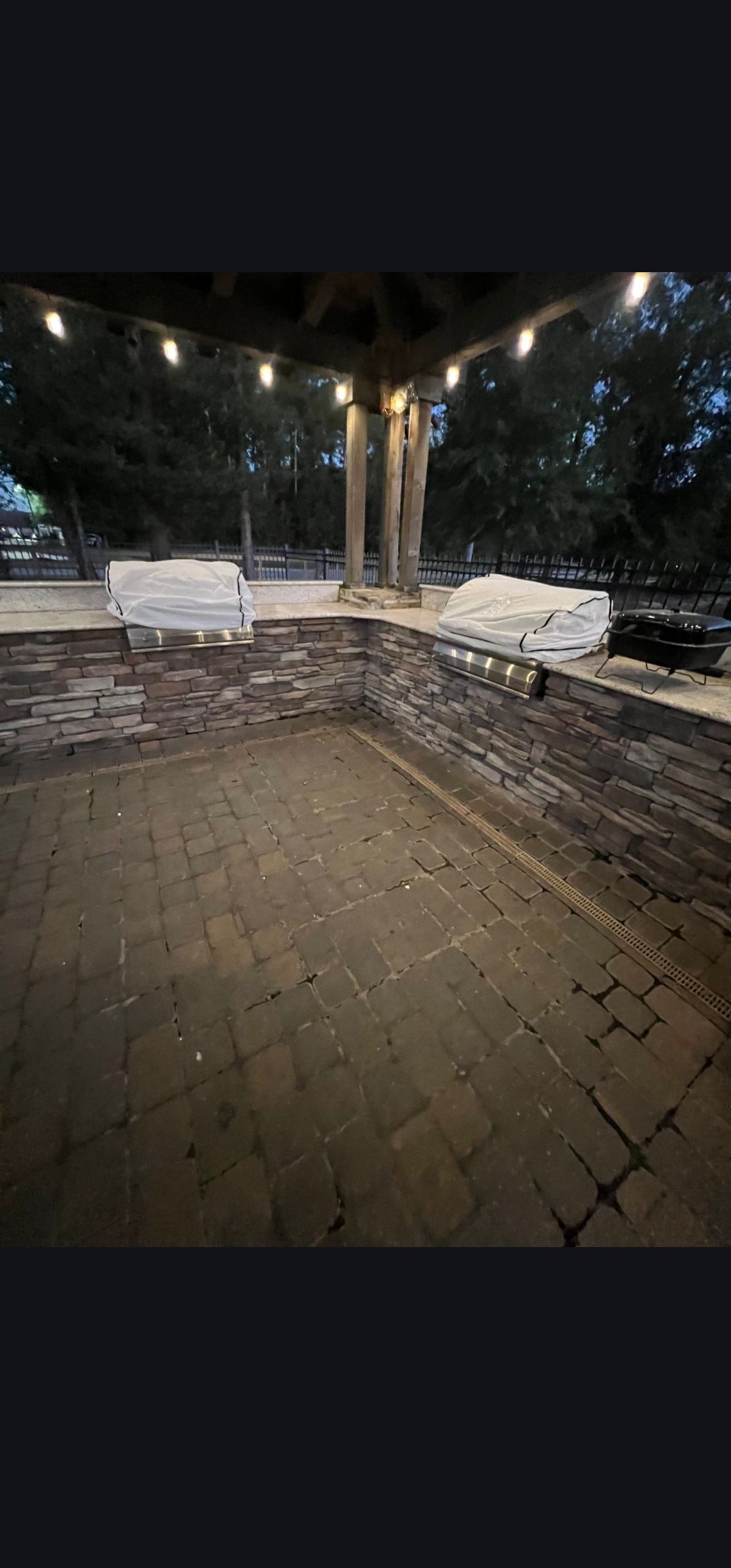 Empty covered patio at night with wet brick floor, benches, and trees lit by streetlights