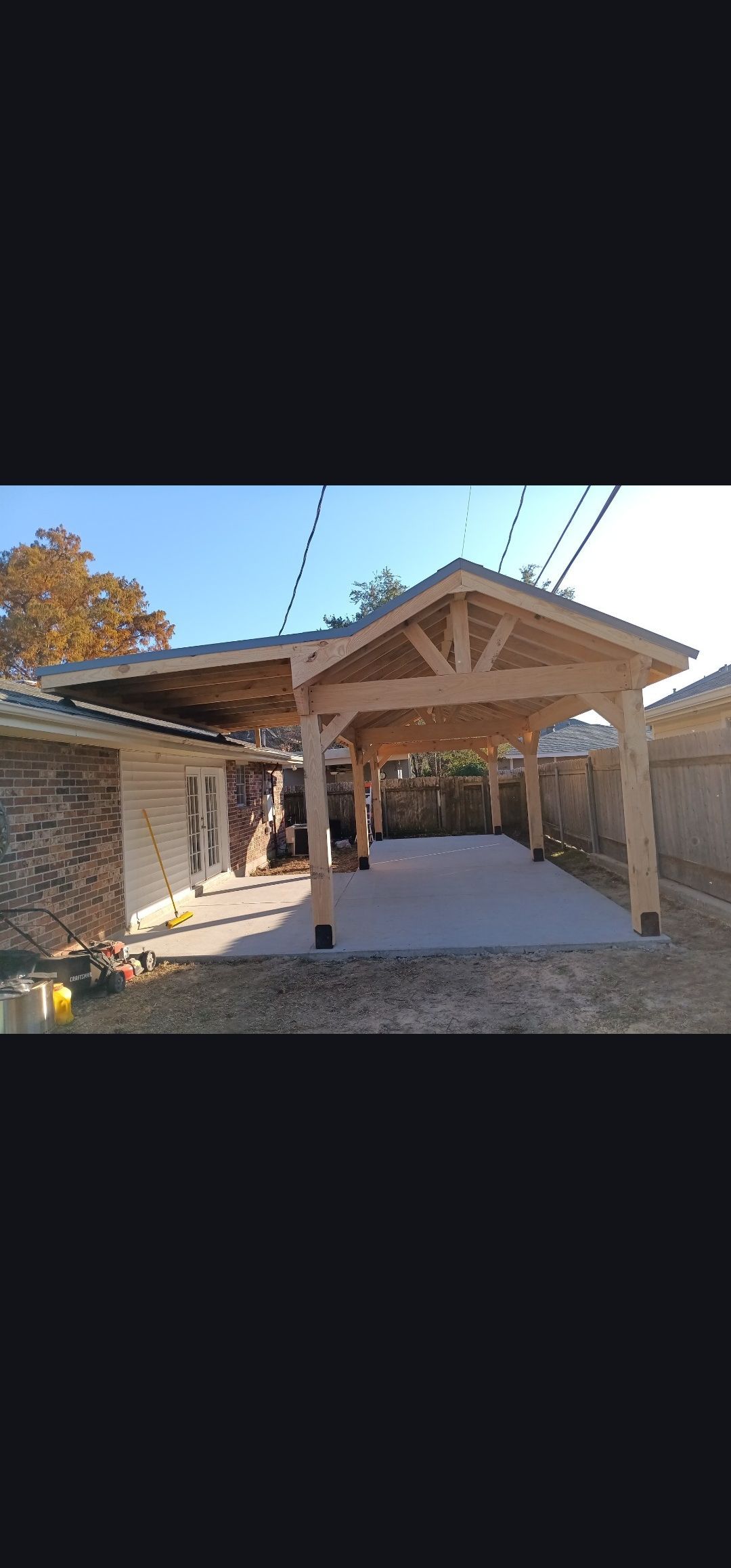 Wooden pavilion under construction with a peaked roof and gravel ground on a sunny day.