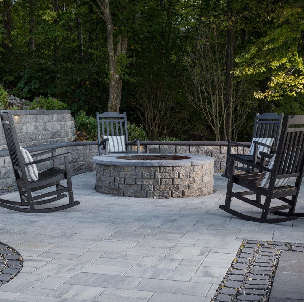 Stone fire pit with four black rocking chairs on a paved patio surrounded by trees and stone seating.