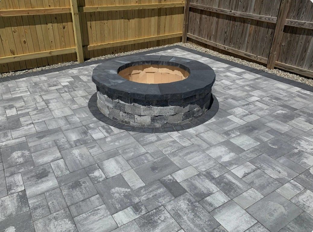 Stone fire pit in a paved backyard surrounded by wooden fencing.