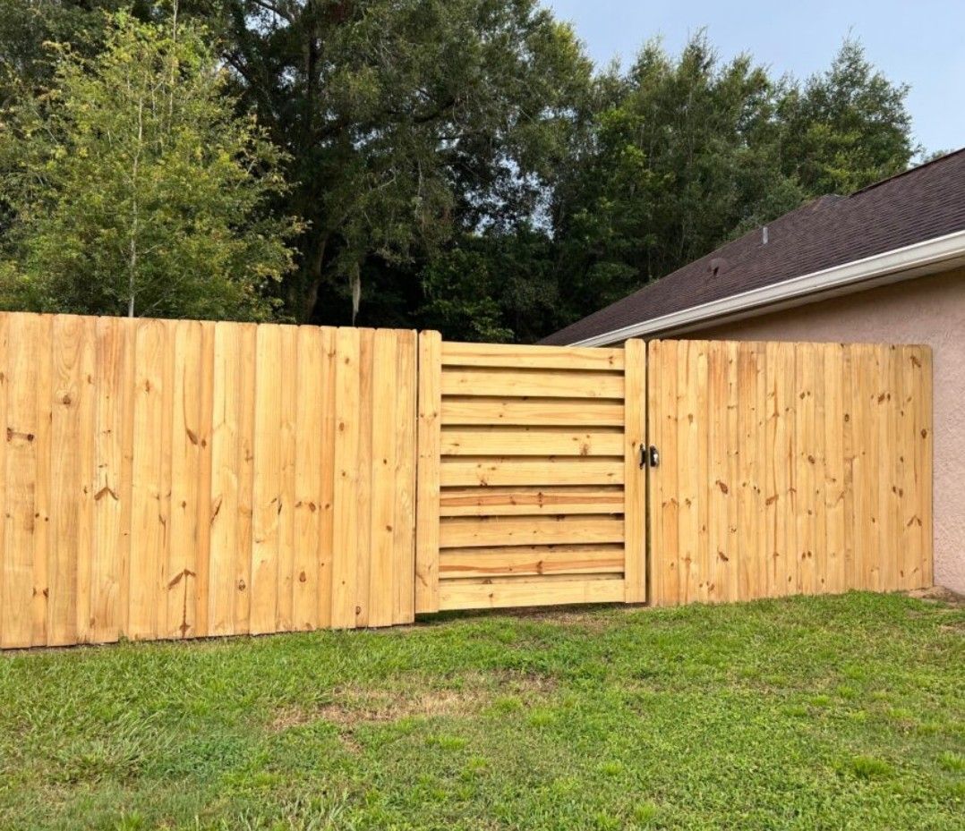 Wooden privacy fence with centered gate in a grassy backyard, bordered by trees and a house roof.