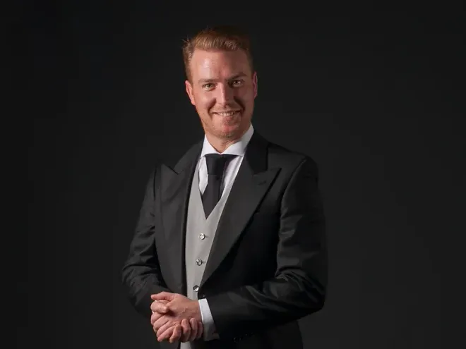 Een man in een smoking en stropdas staat met zijn handen gevouwen en glimlacht.