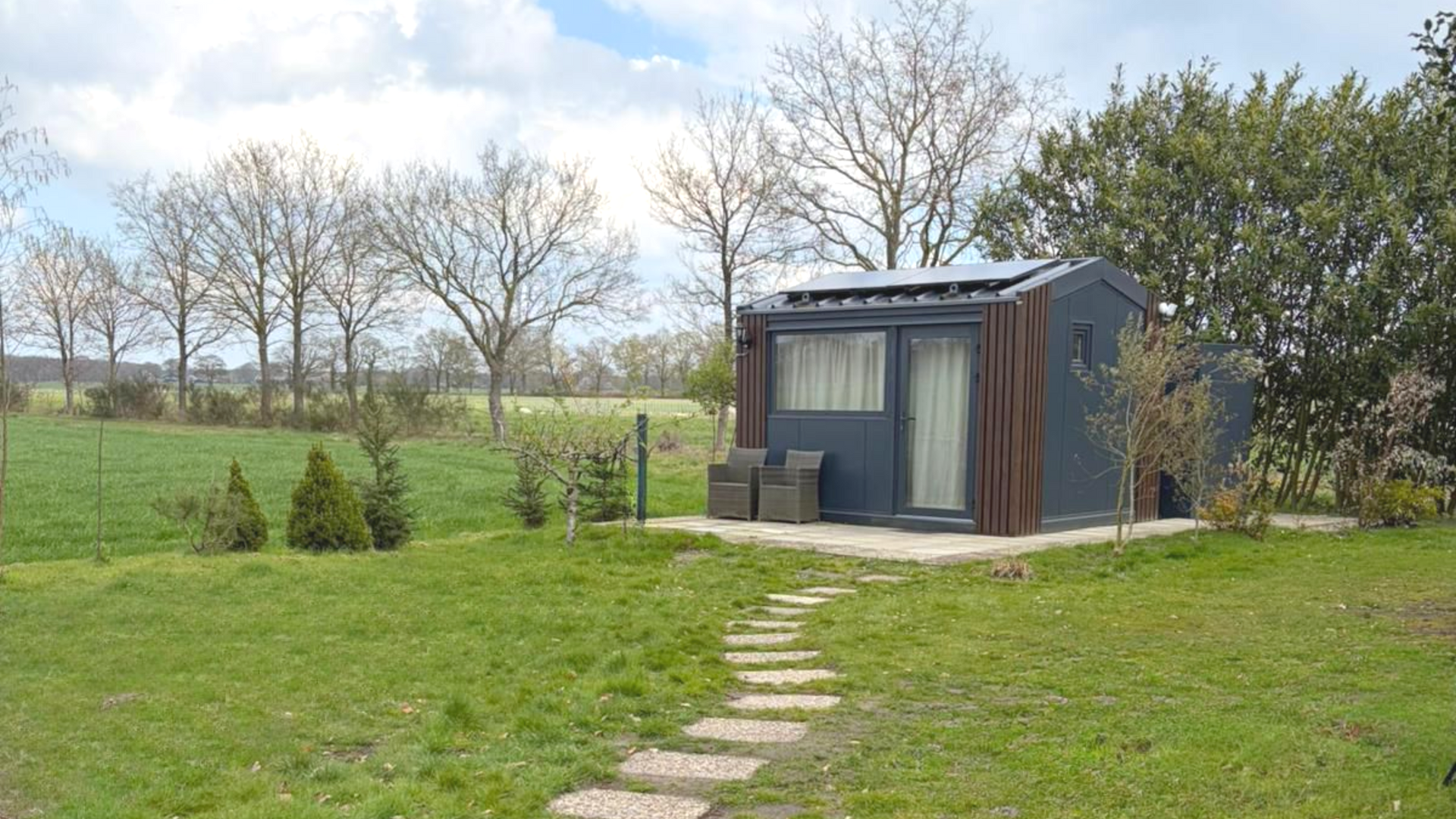 Een klein, donker schuurtje met een stenen pad op een grasveld, omgeven door kale bomen en open velden.