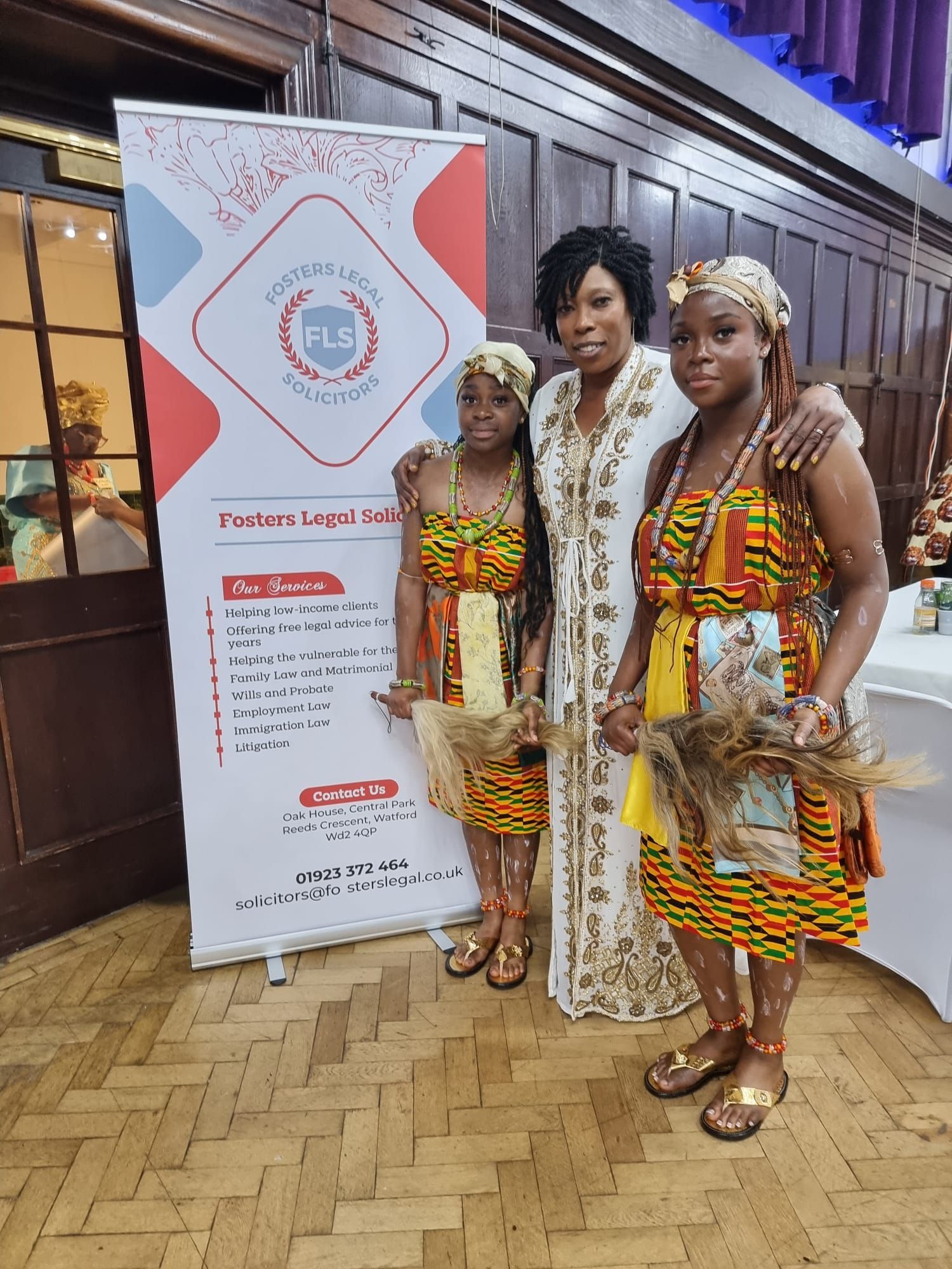 Three people pose for a photo near a banner. Two young women in traditional African dress stand on either side of a woman wearing a white embroidered dress. They are indoors.