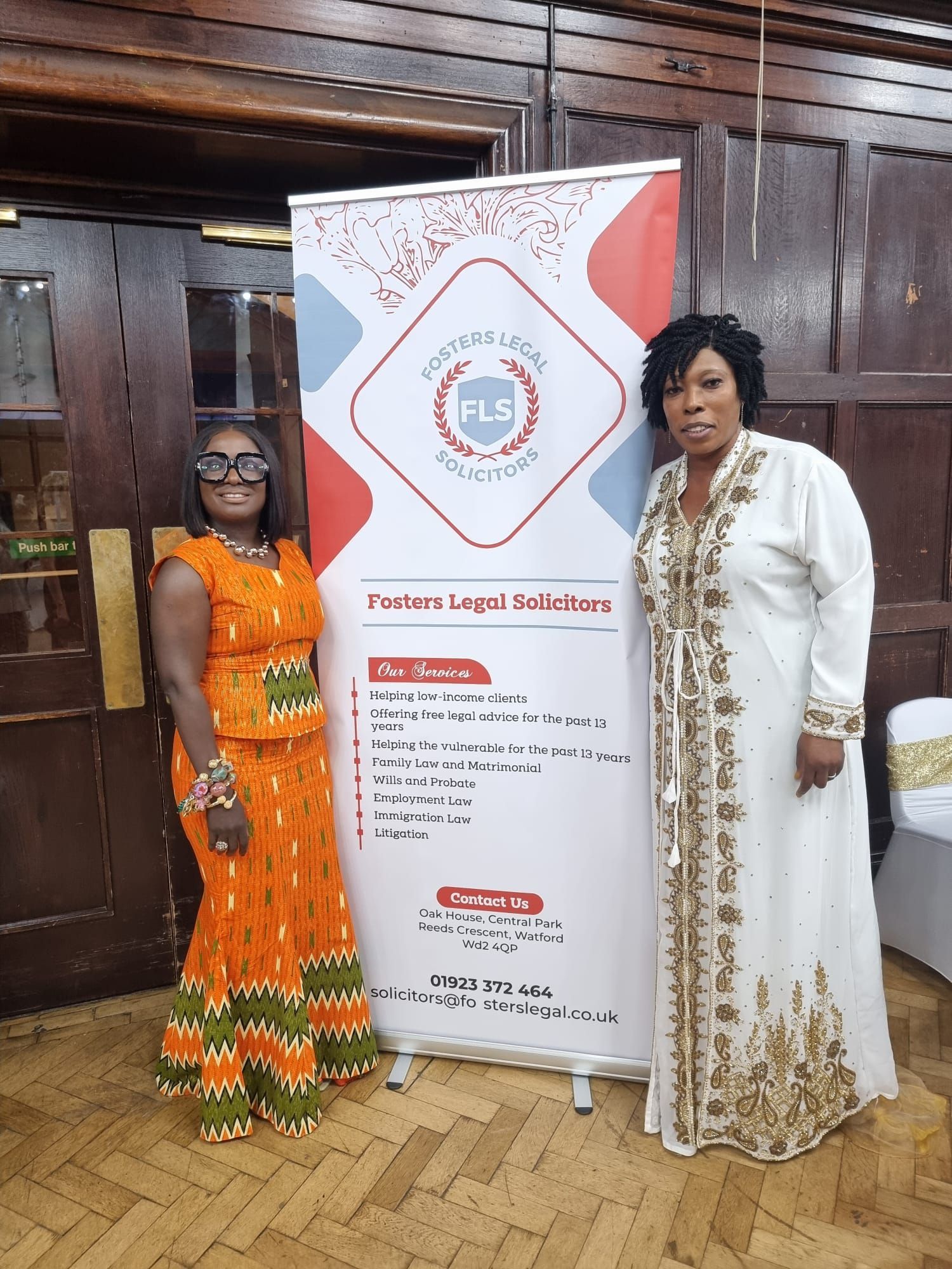 Two women stand next to a banner in a wood-paneled room. One wears orange and the other a white gown. The banner features a logo and text.