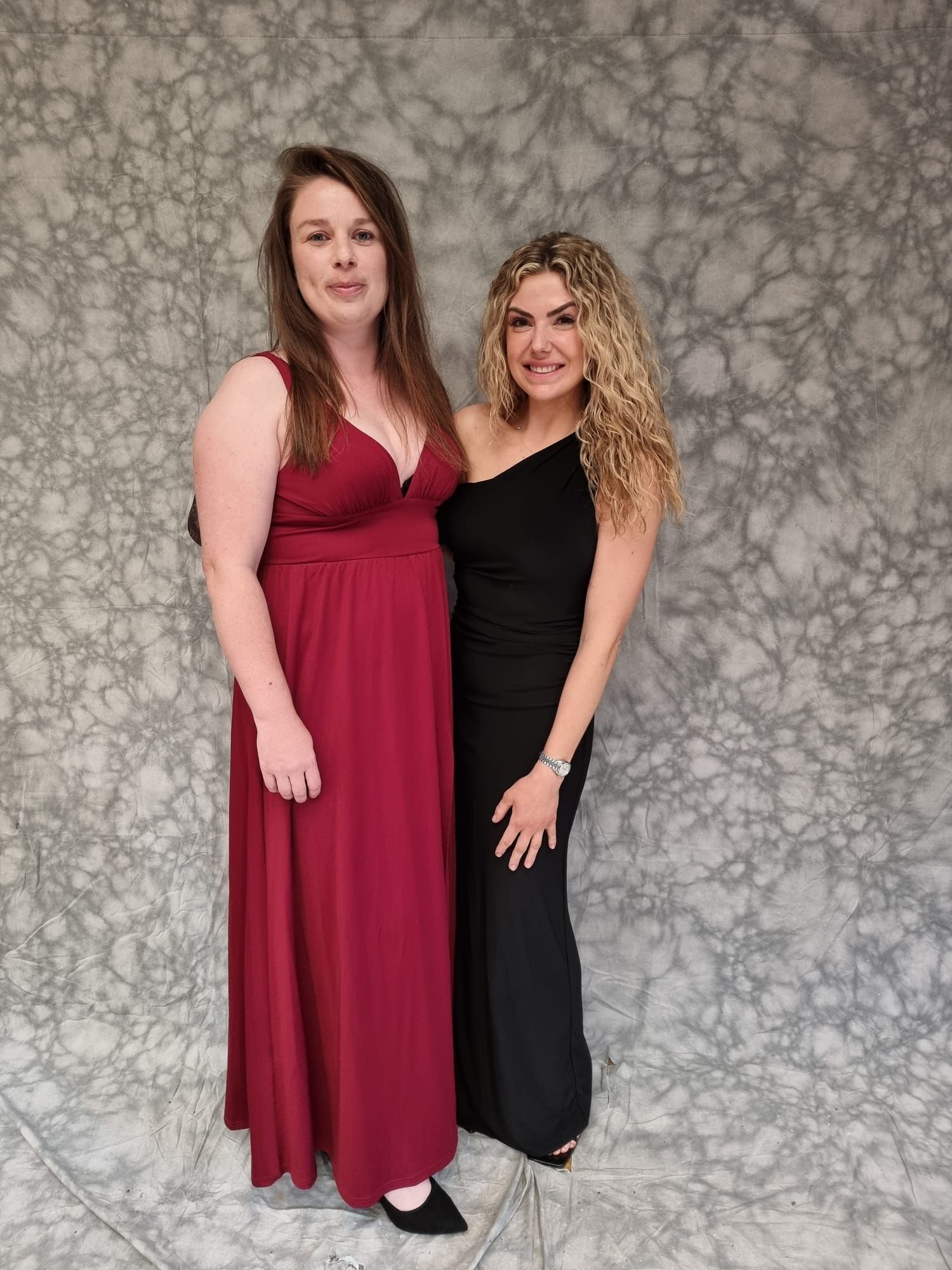 Two women in formal dresses pose in front of a mottled gray backdrop. One wears red with a V-neck, the other a black one-shoulder.